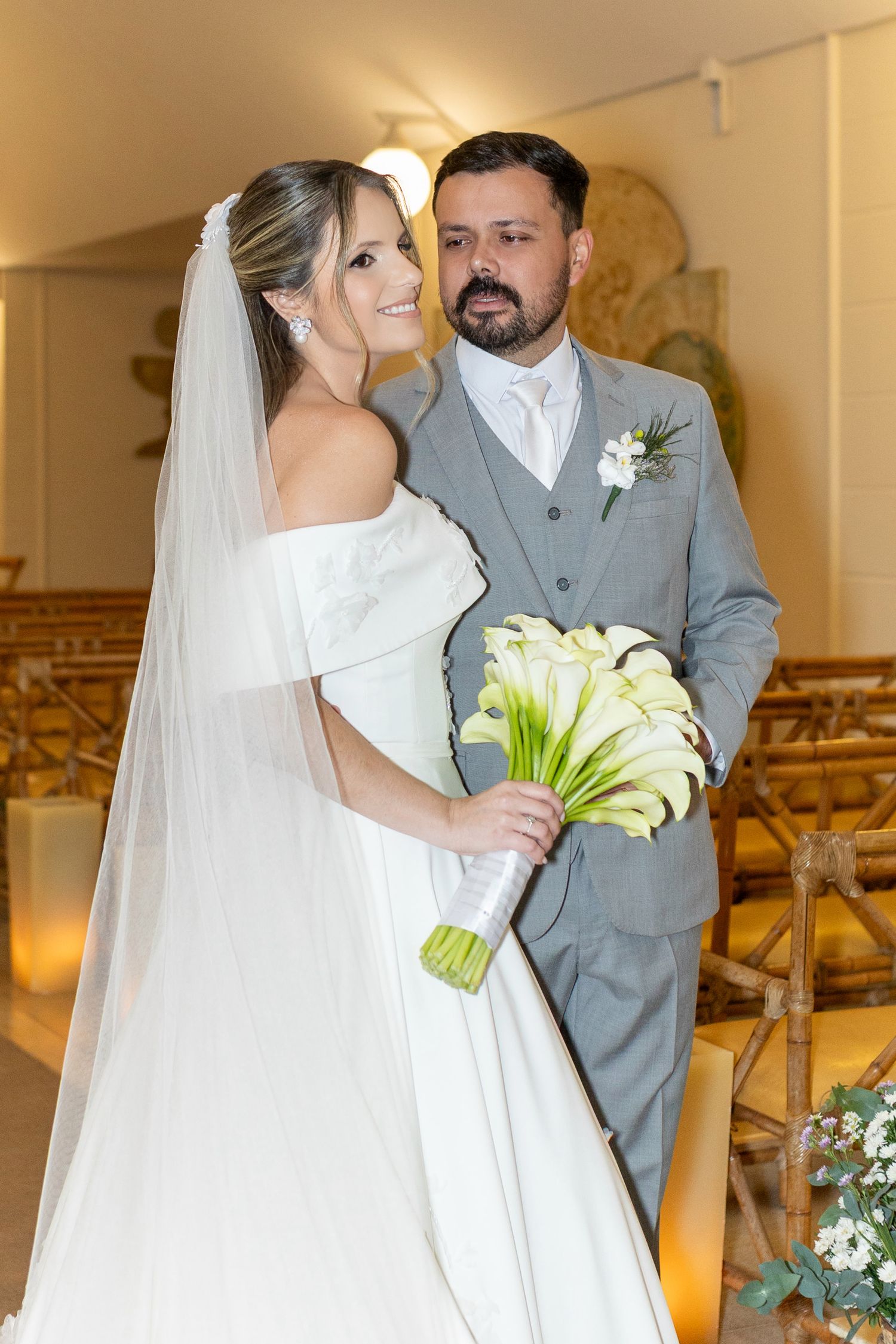 Bride in white off-shoulder gown with veil holds calla lily bouquet beside groom in gray suit inside a church.