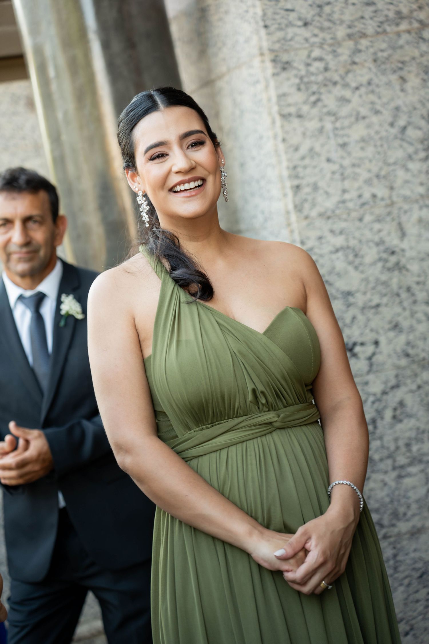 Smiling woman in elegant olive green one-shoulder gown with drop earrings at a wedding event.