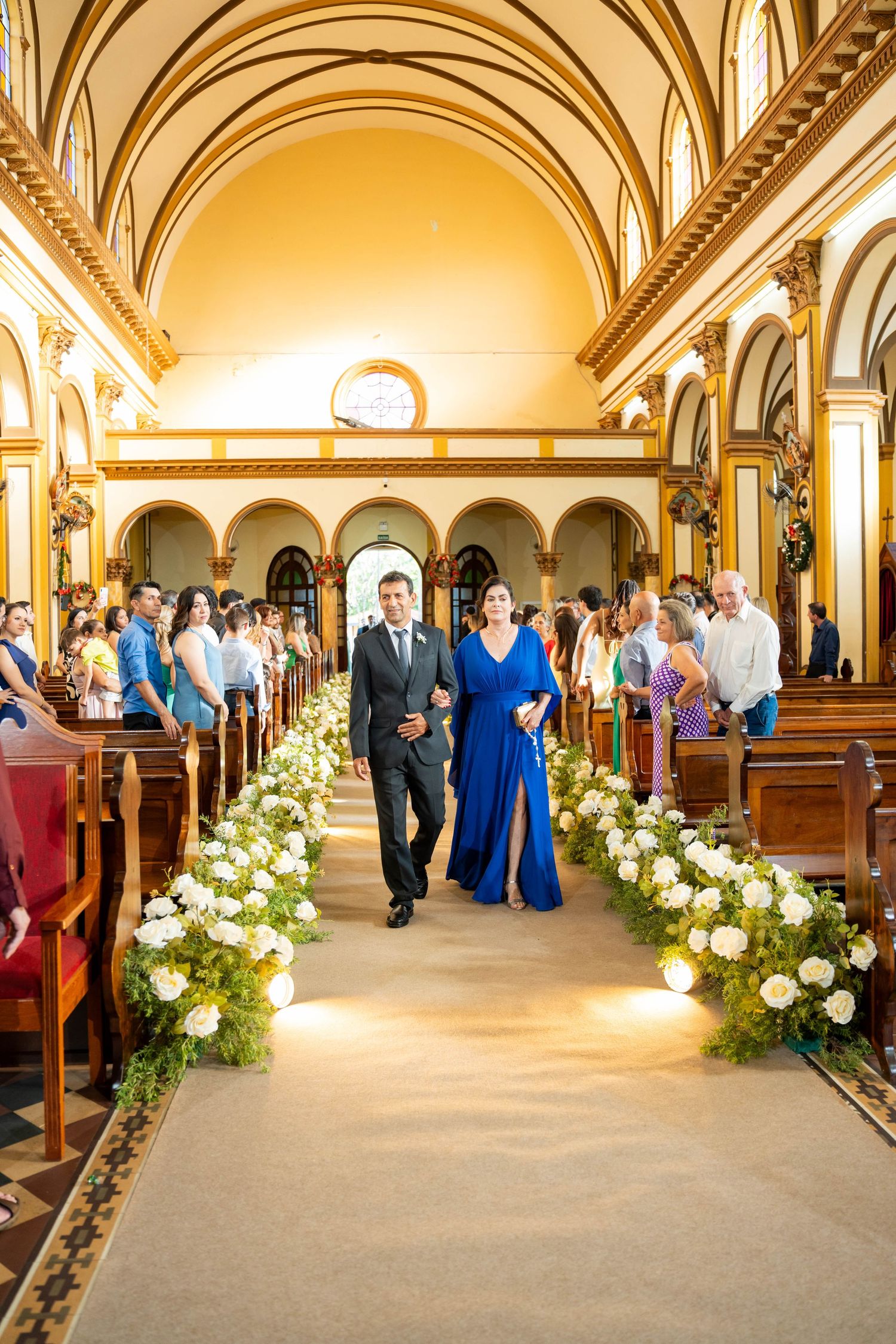 Elegant church wedding ceremony with couple walking down floral aisle lined with white roses in golden ornate chapel.