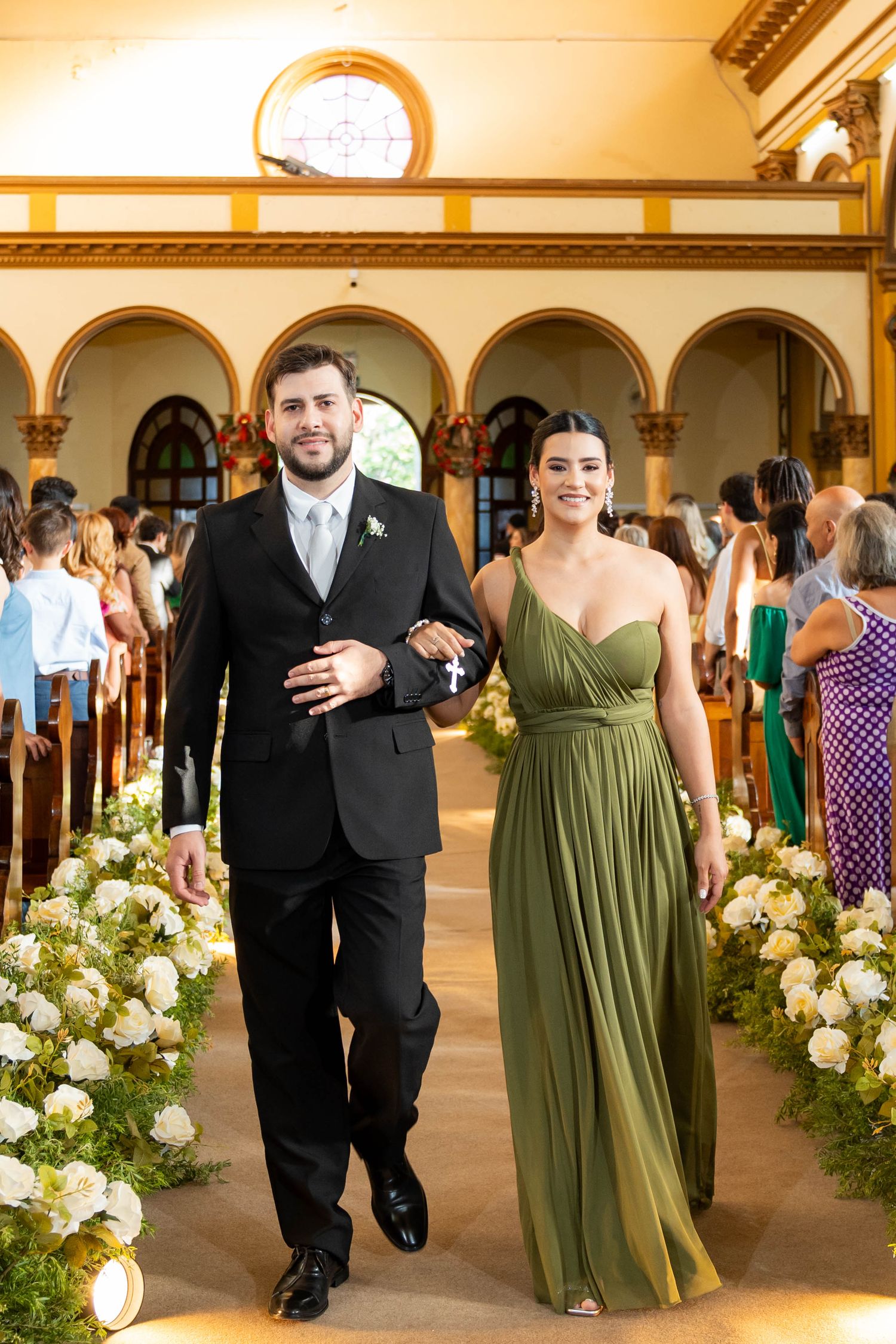 A groomsman in a black tuxedo escorts a bridesmaid in an olive green gown down a floral-lined church aisle.