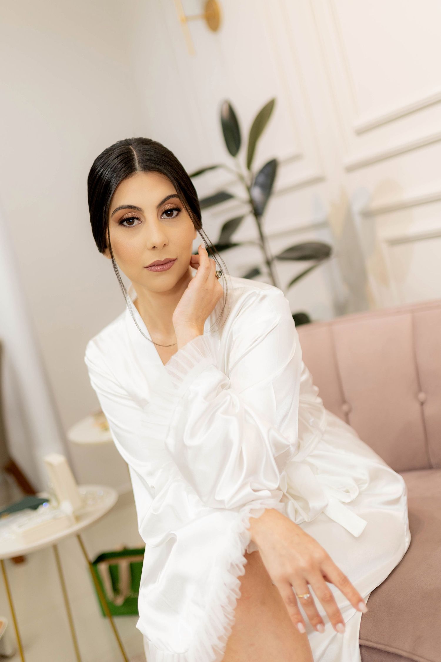 Bride in white satin robe sitting elegantly, touching her ear, with soft interior decor in background.