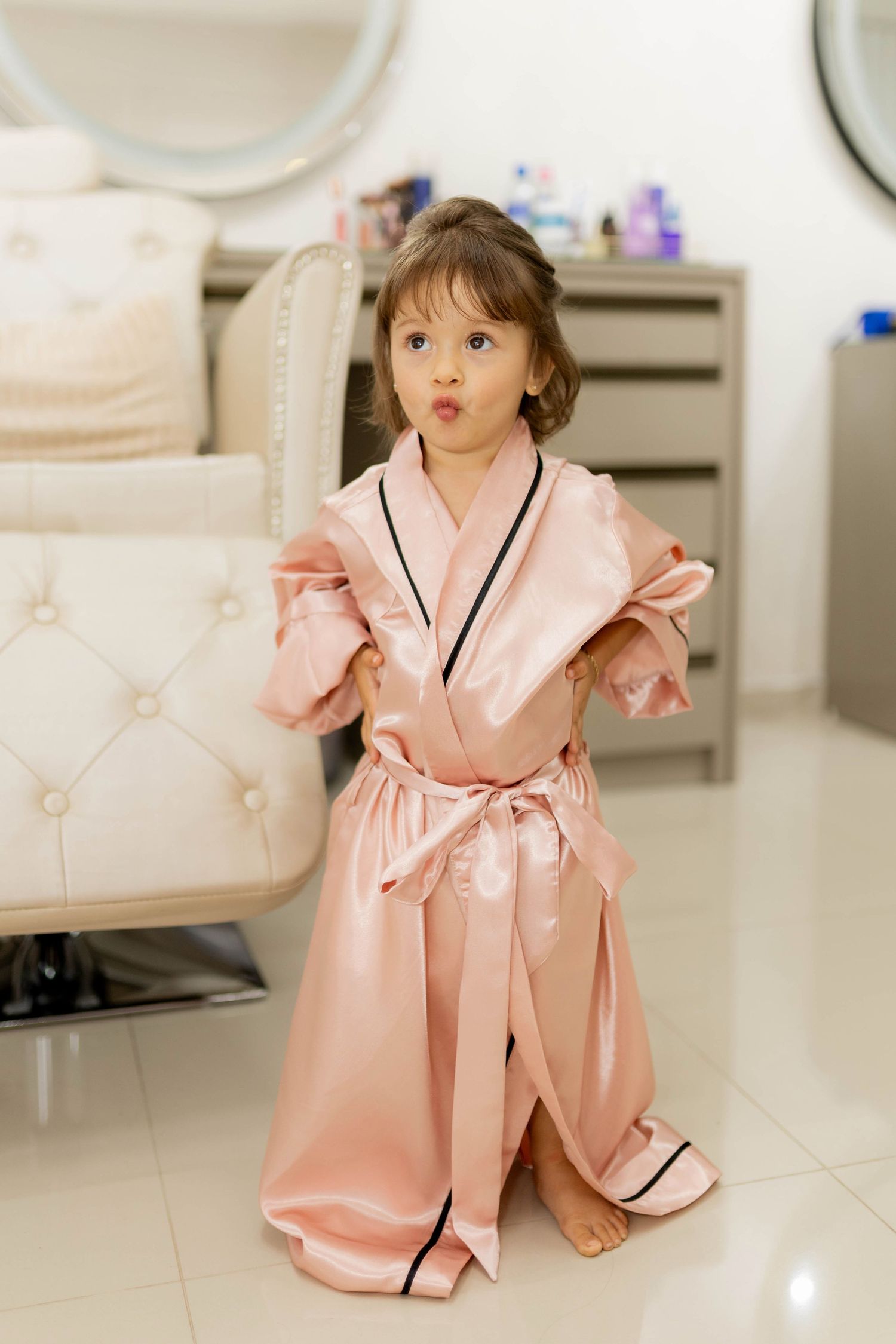 Adorable little girl wearing an oversized pink satin robe with black trim, posing in a beauty salon.