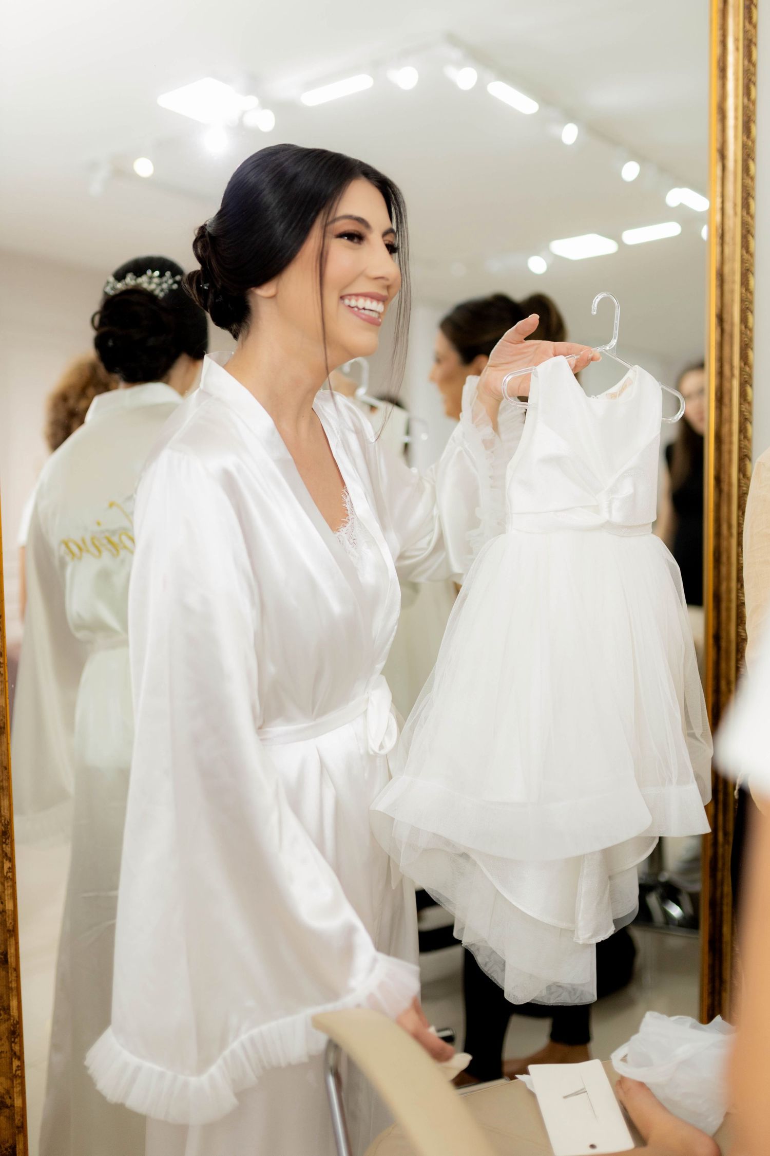 Bride in white robe smiling while holding a flower girl dress on a hanger in a bridal suite.