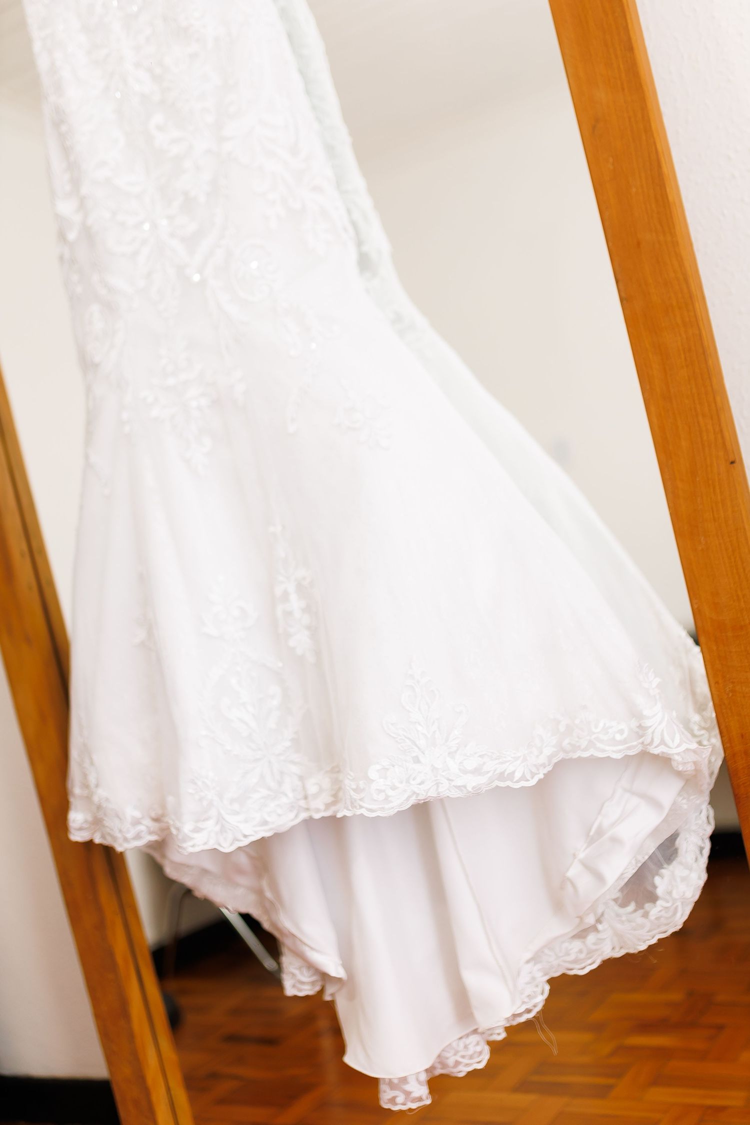 White lace mermaid wedding dress hanging on a wooden door frame, featuring intricate floral embroidery and scalloped hem.