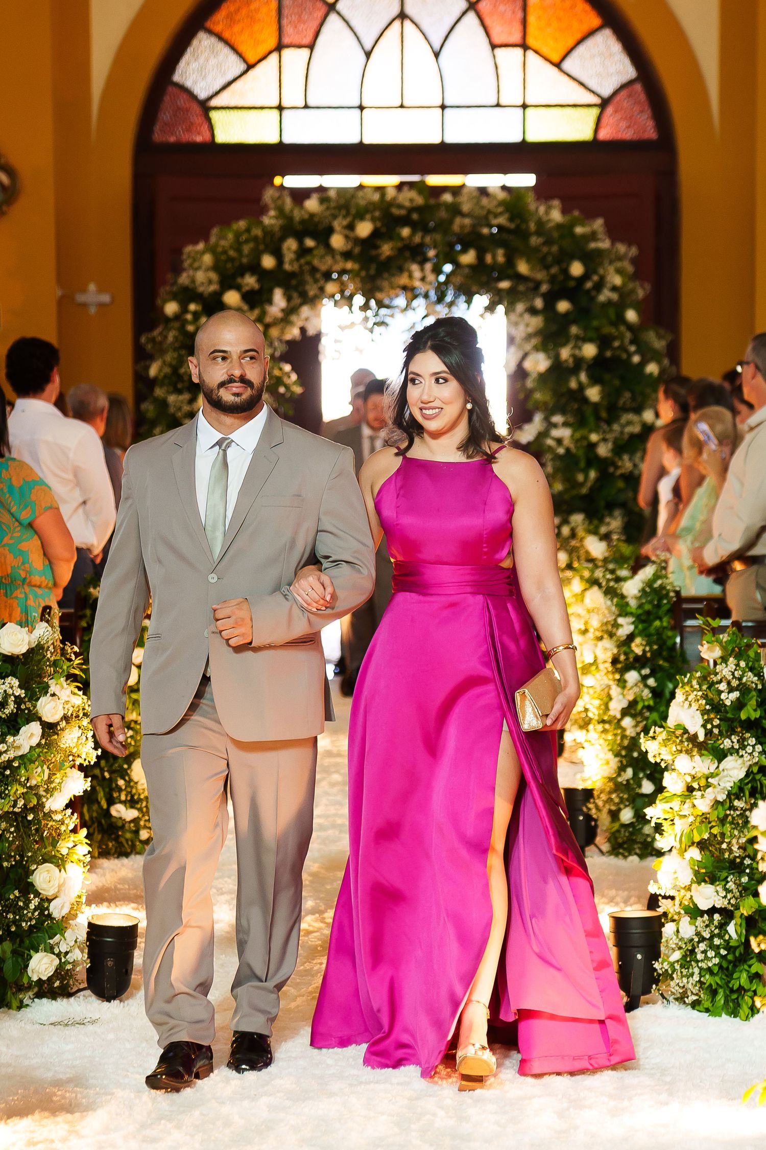 Couple walking down wedding aisle, man in tan suit, woman in elegant fuchsia gown, floral arch backdrop.