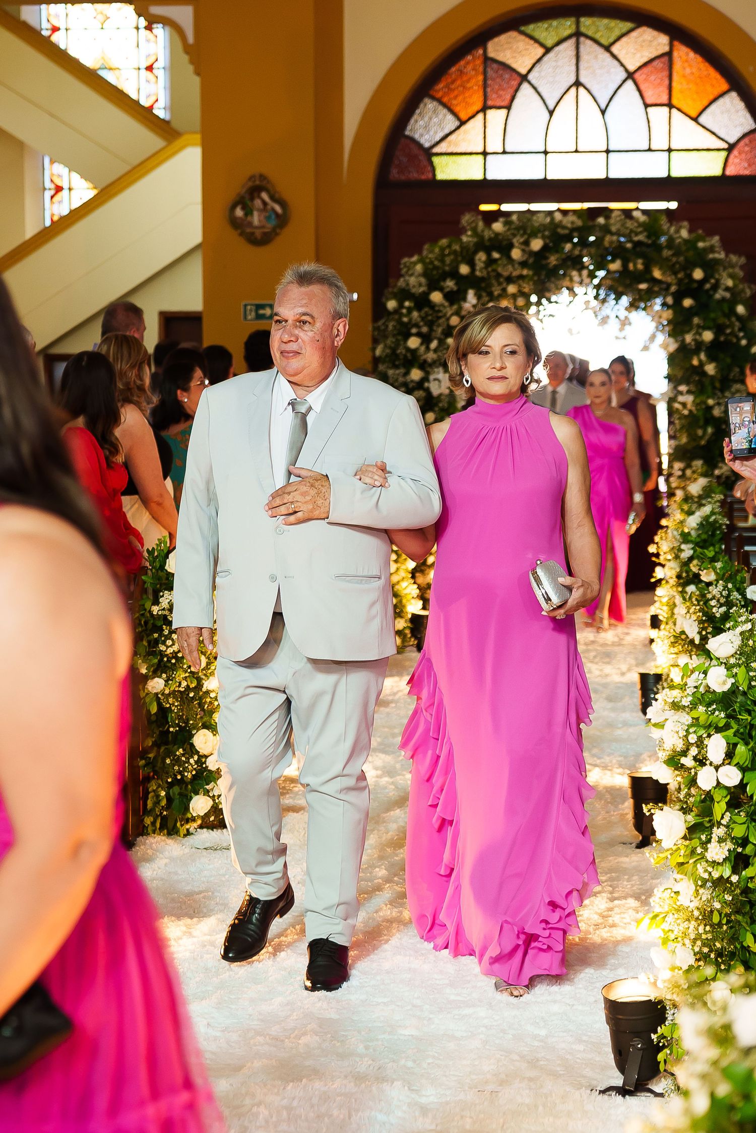 Elegant couple walking down floral wedding aisle, man in gray suit, woman in pink gown, church ceremony.