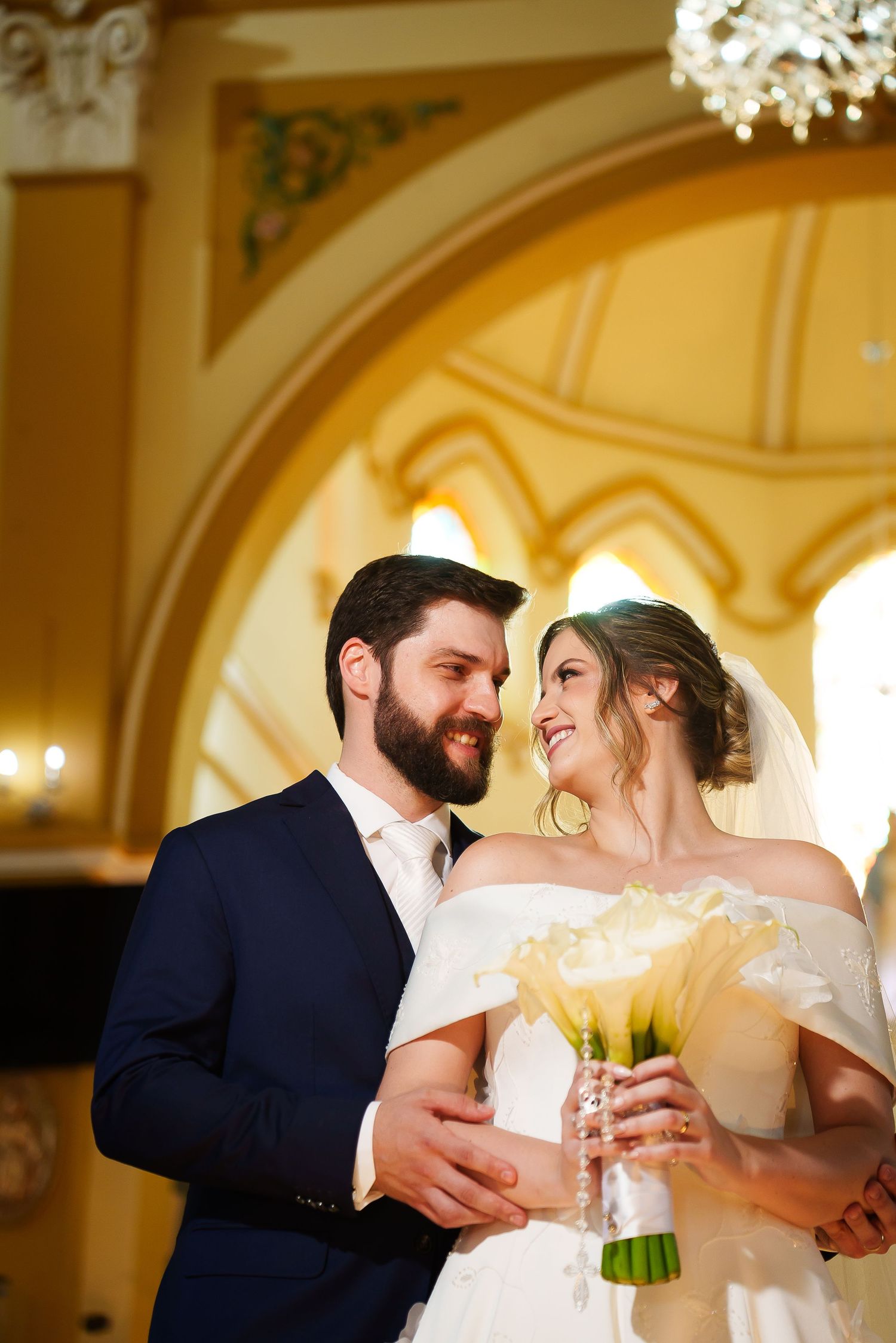 Smiling bride and groom embrace in elegant church, bride holds white calla lily bouquet wearing off-shoulder gown.