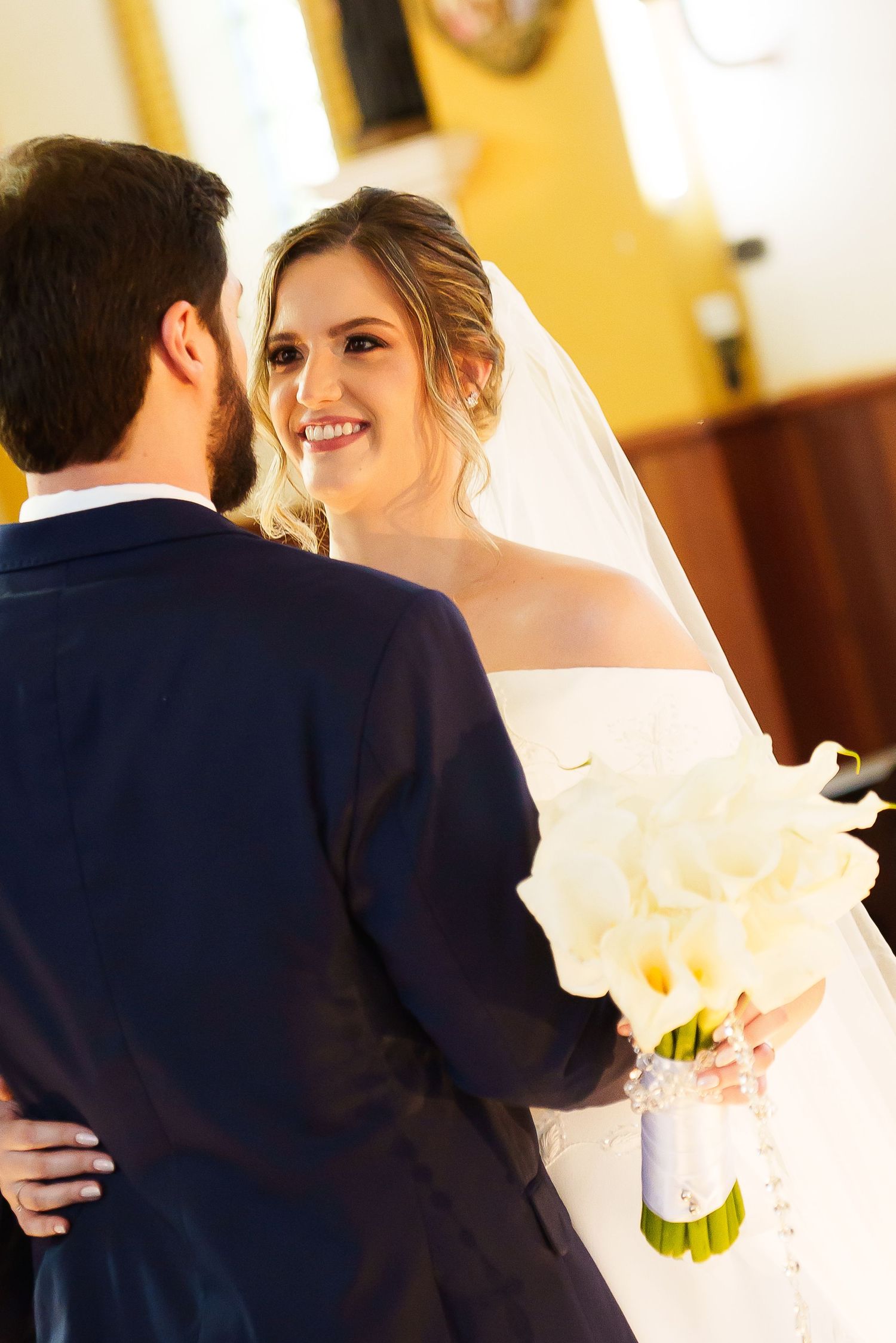Bride smiling at groom during wedding ceremony, holding white calla lily bouquet, wearing veil and off-shoulder gown.