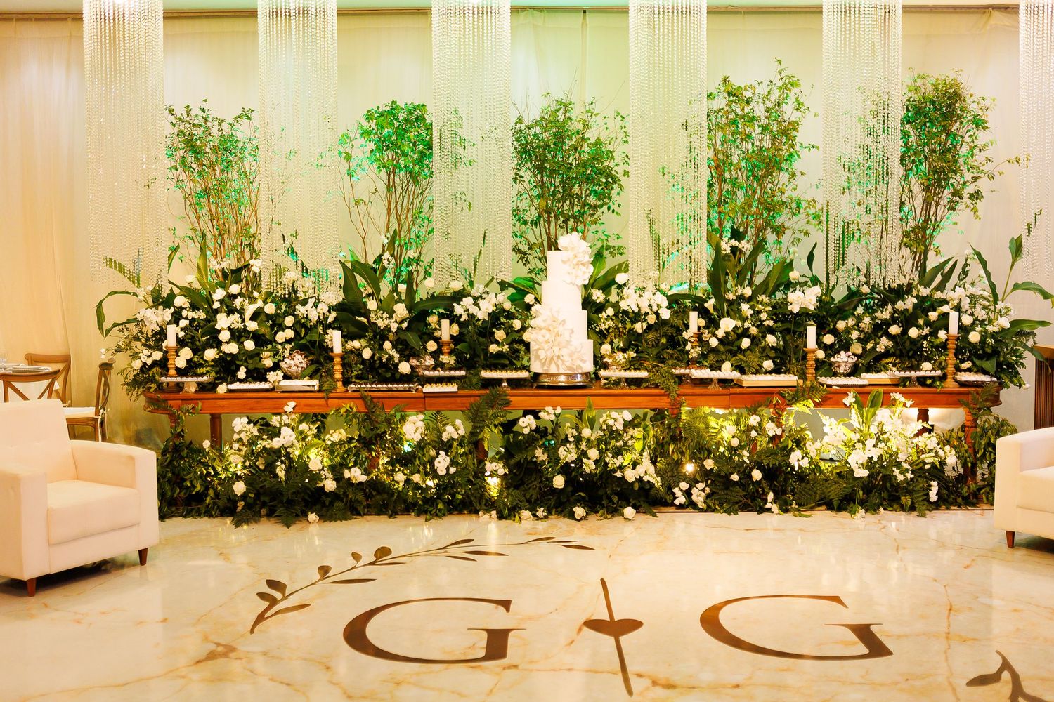 Elegant wedding reception table adorned with lush greenery, white floral arrangements, and a tiered cake against a backdrop of tall trees.