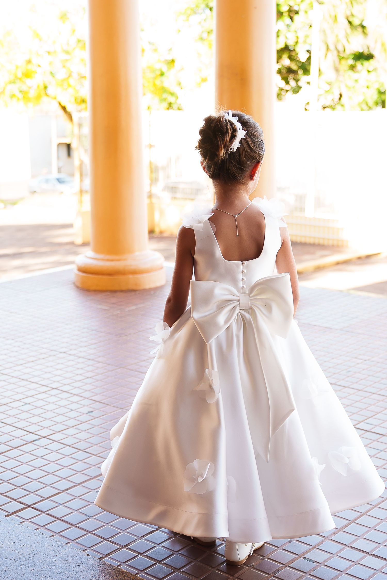 Young girl in white flower girl dress with large bow, viewed from behind, standing between yellow columns.