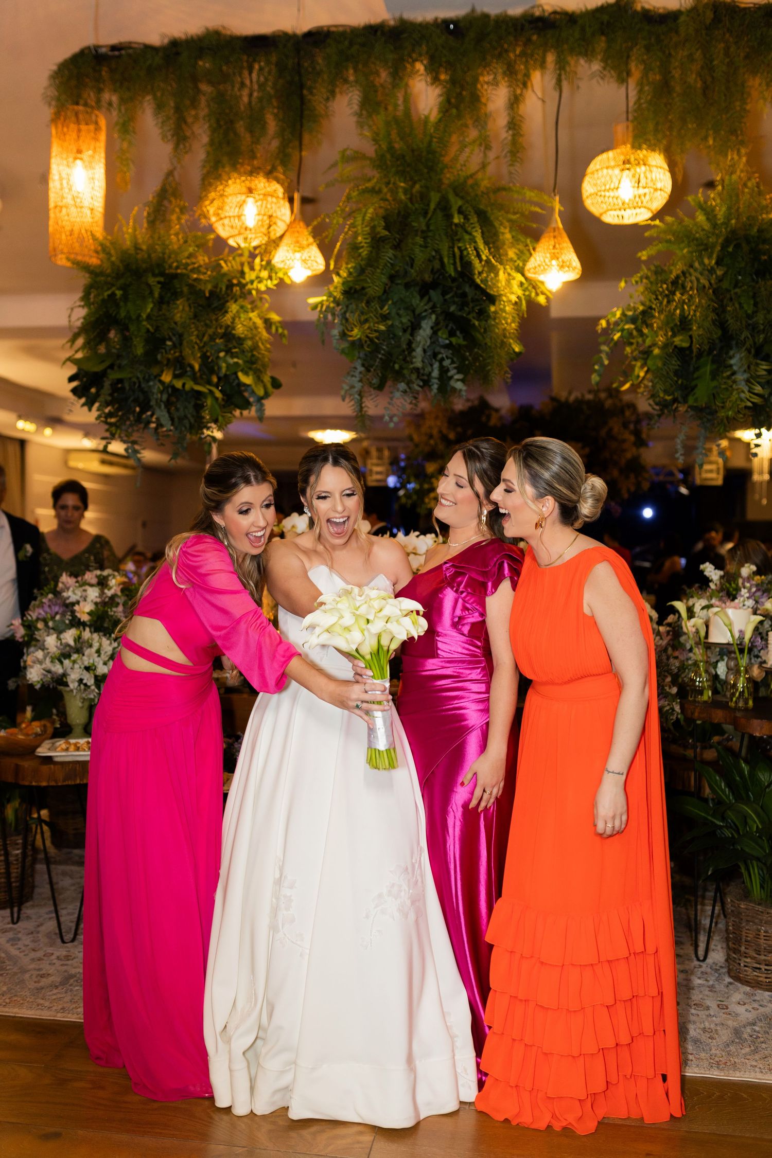 Bride in white gown holds calla lily bouquet surrounded by bridesmaids in pink and orange dresses at elegant wedding.