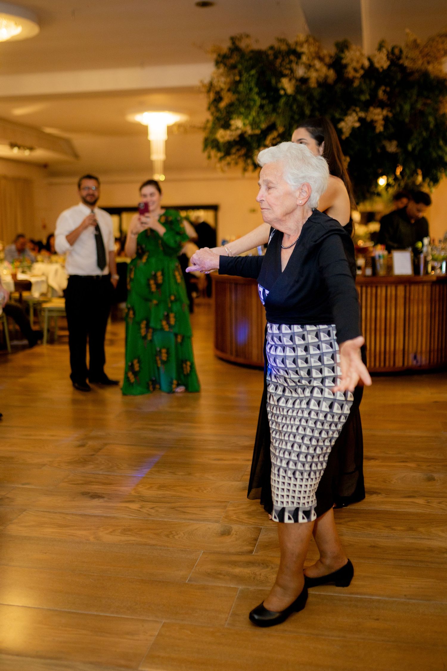 Elderly woman dancing gracefully at a wedding reception in a patterned skirt and black top on a wooden dance floor.
