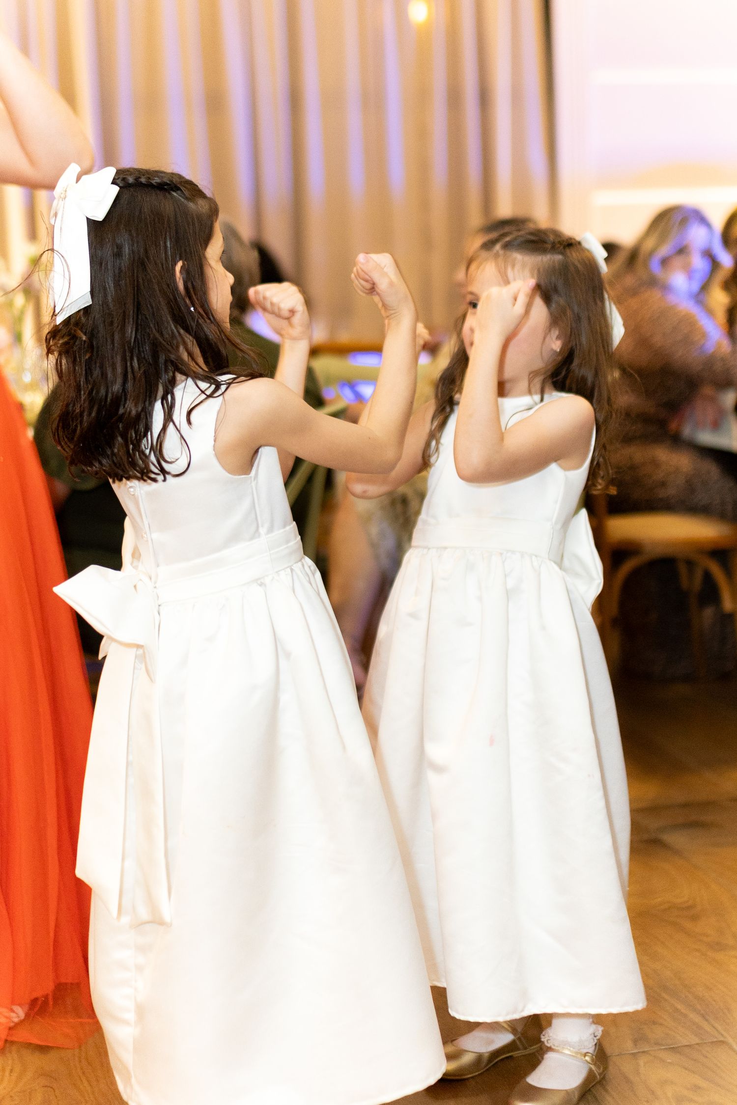 Two young flower girls in white dresses dancing together at a wedding reception.