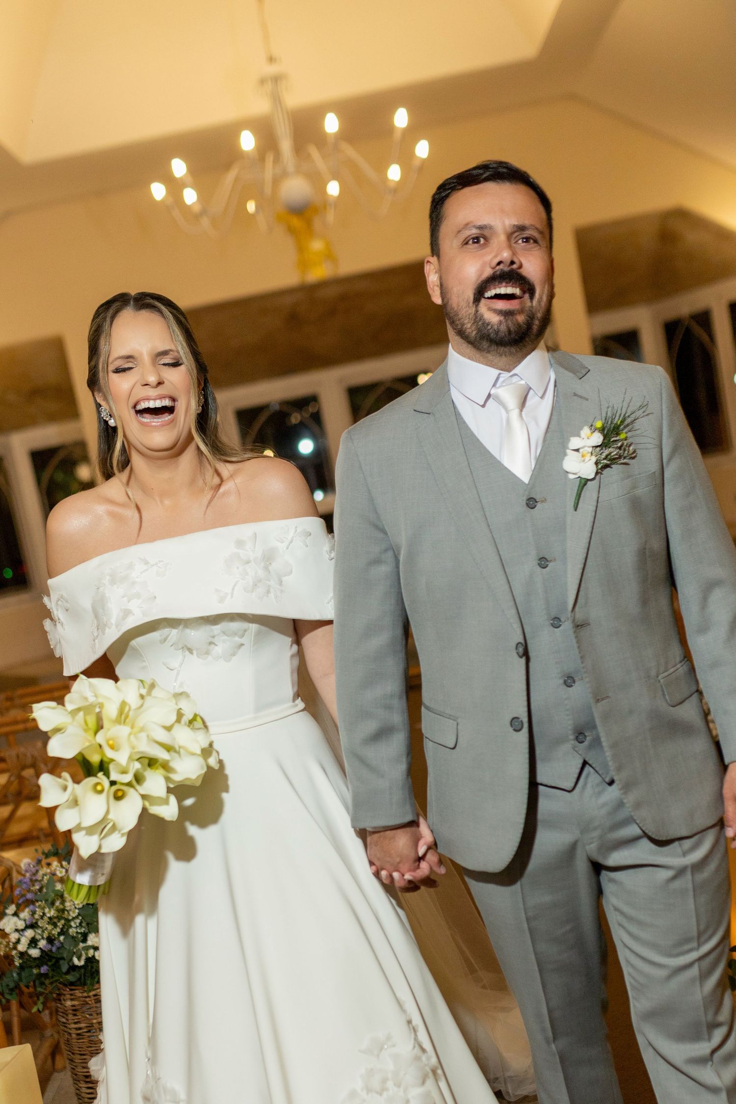Joyful bride in white off-shoulder gown holding calla lily bouquet and groom in gray suit laughing together at wedding reception.