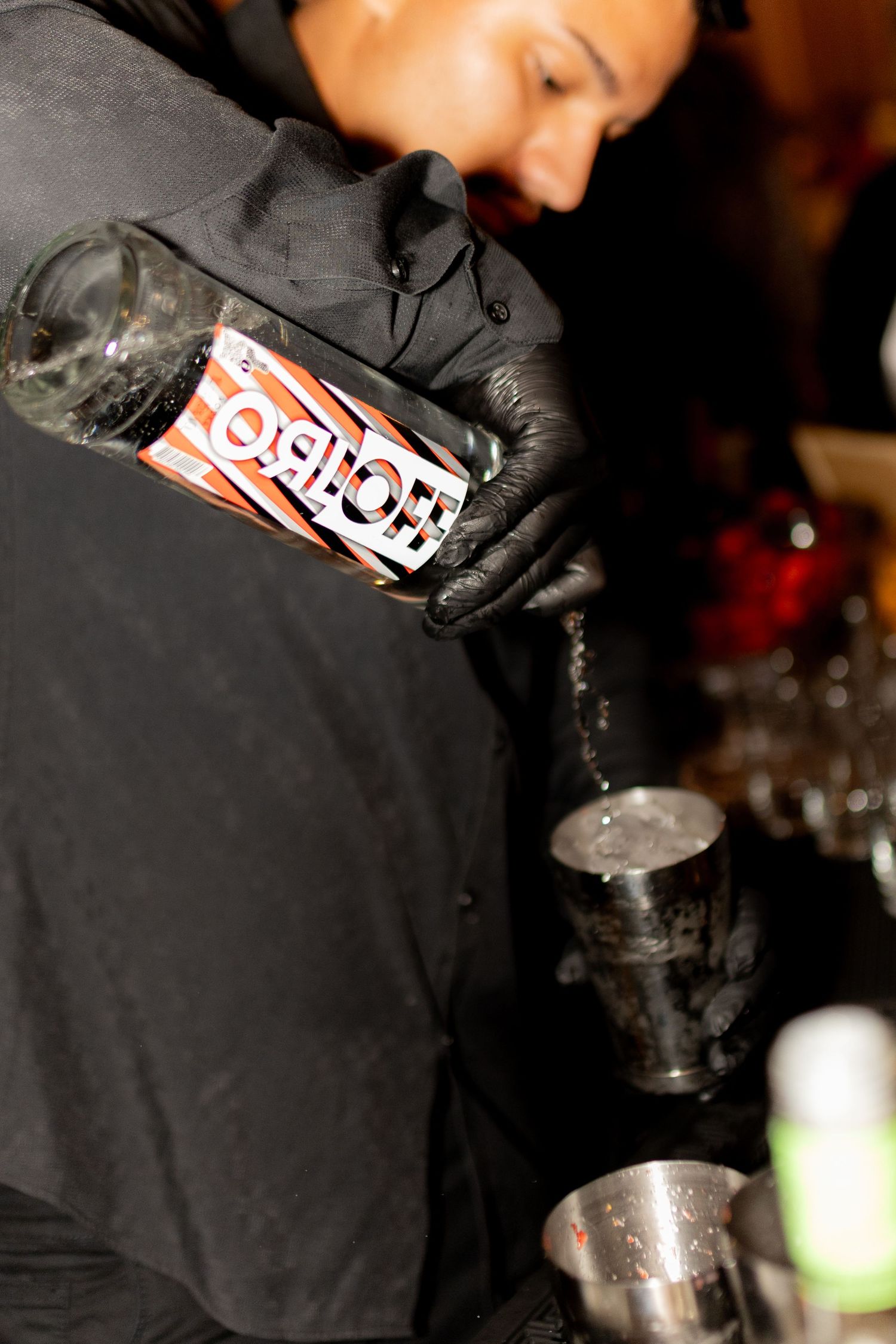 Bartender in black gloves pouring a can of Fanta into a jigger while mixing cocktails at a bar.
