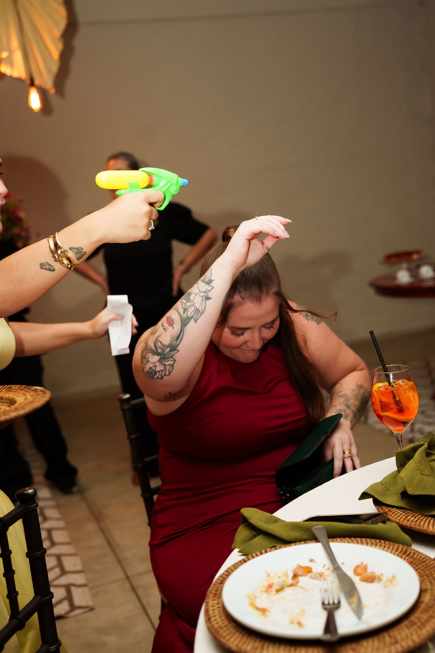 Woman in red dress dodging a colorful water gun at a dinner party, with drinks and plates on the table.