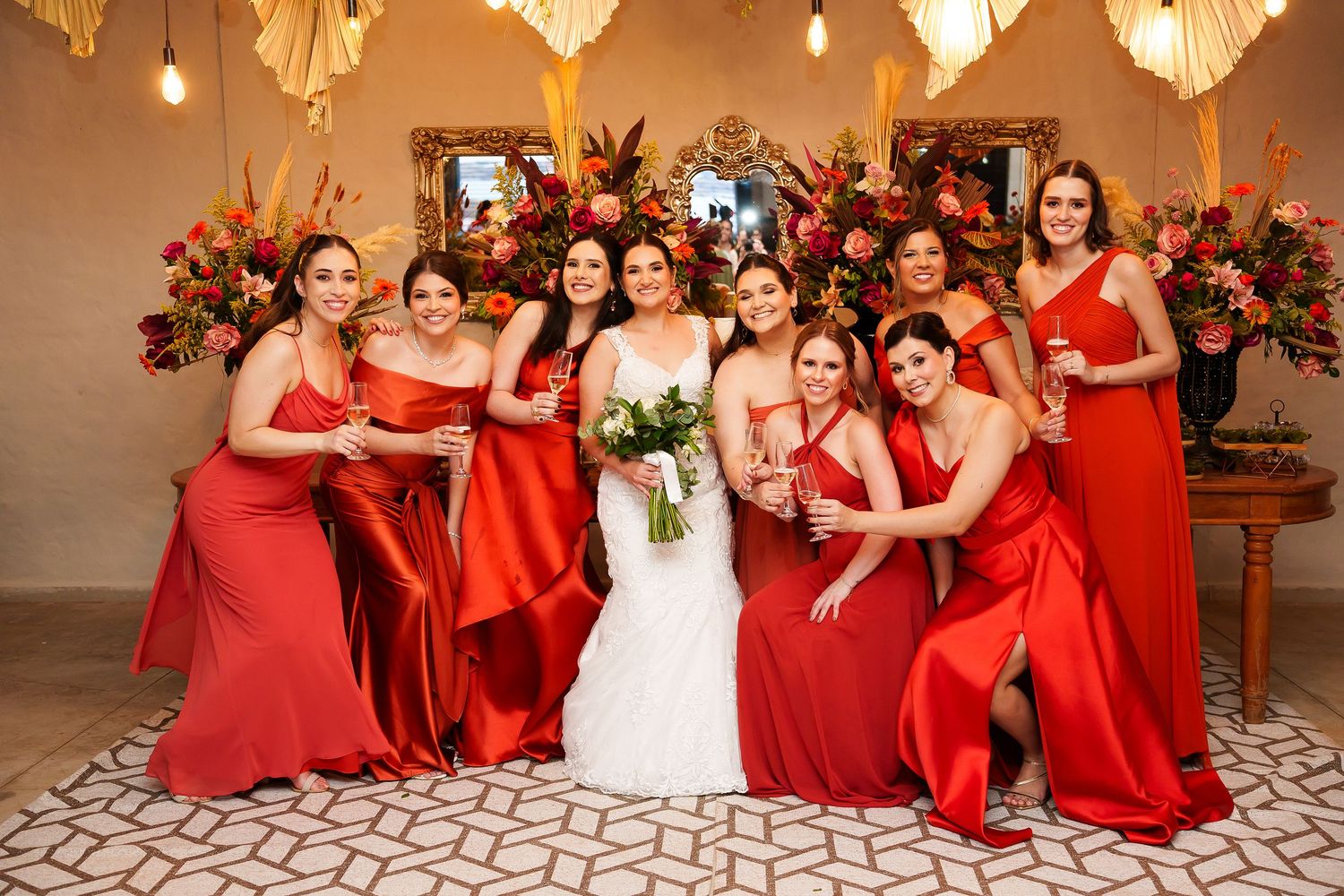 Bride in white gown poses with eight bridesmaids in red satin dresses, holding bouquets in an elegant venue.