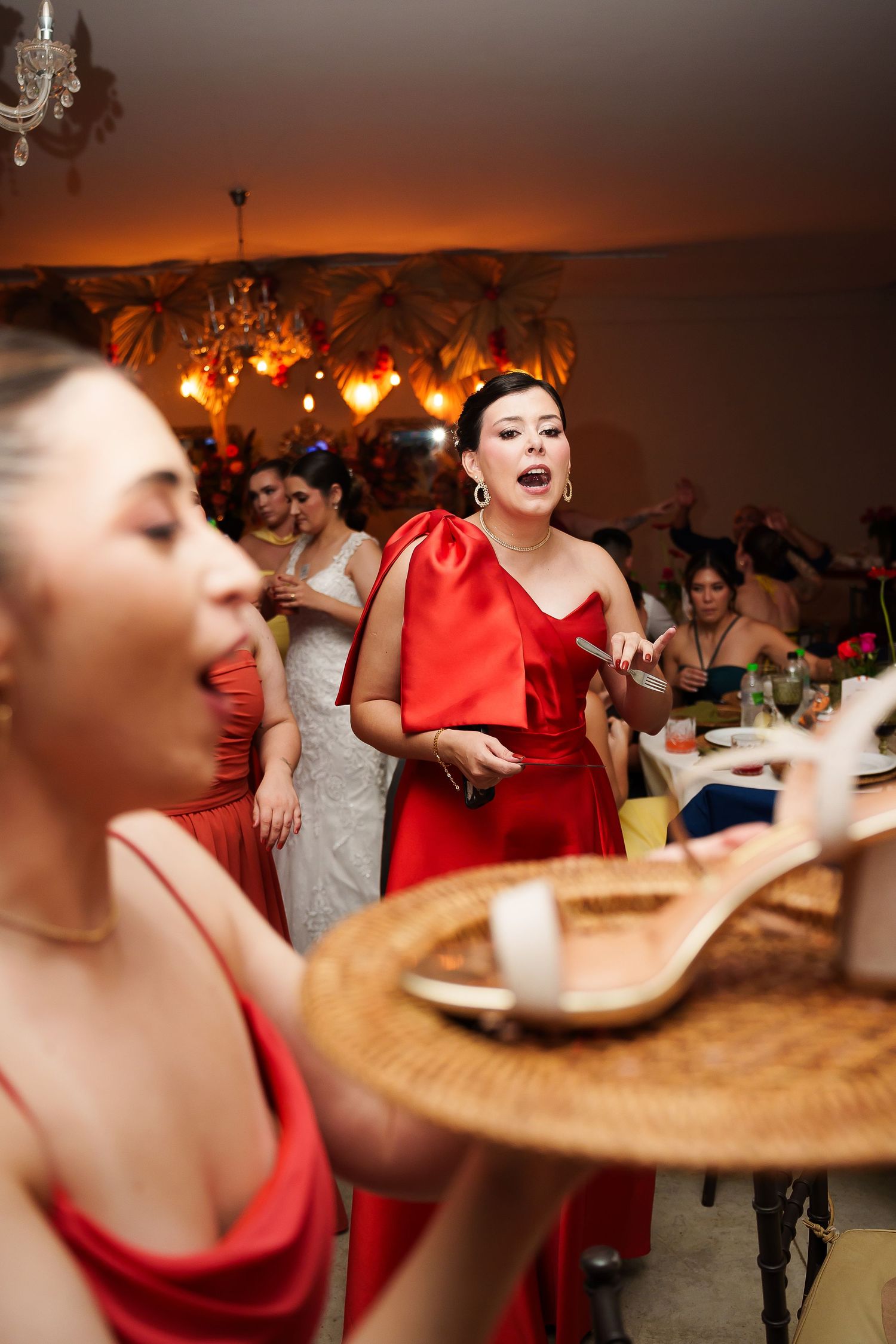 Women in red dresses celebrating at a wedding reception, singing and holding a decorative tray in elegant venue.