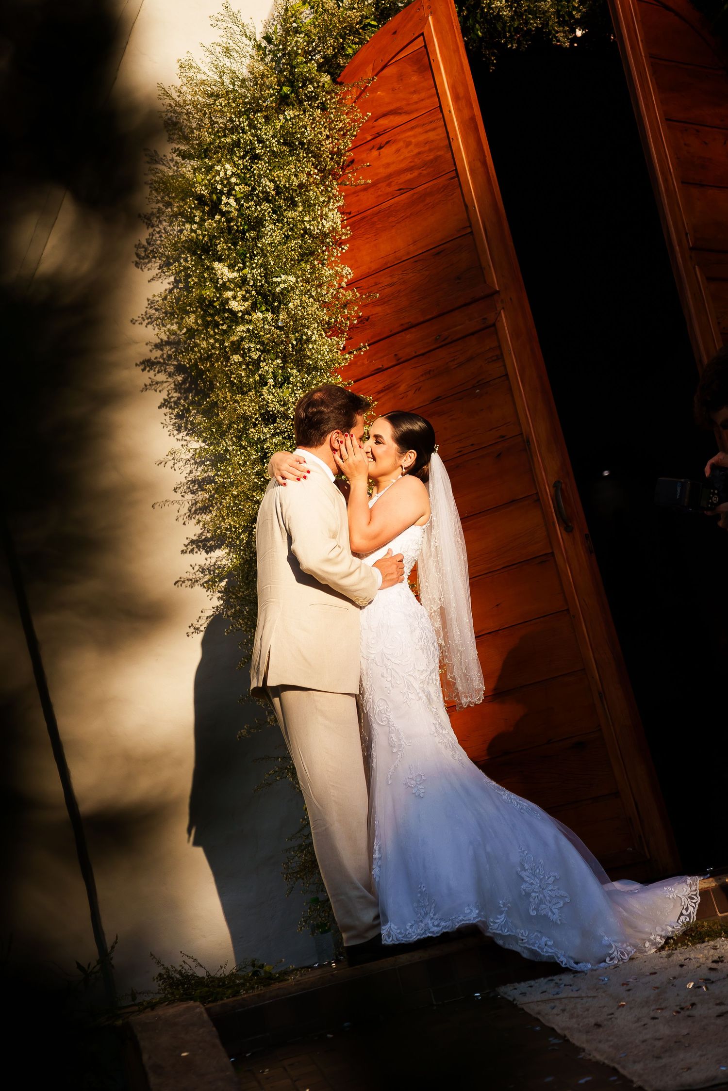 A bride and groom share a romantic kiss near a rustic red wooden door with lush greenery in warm golden light.