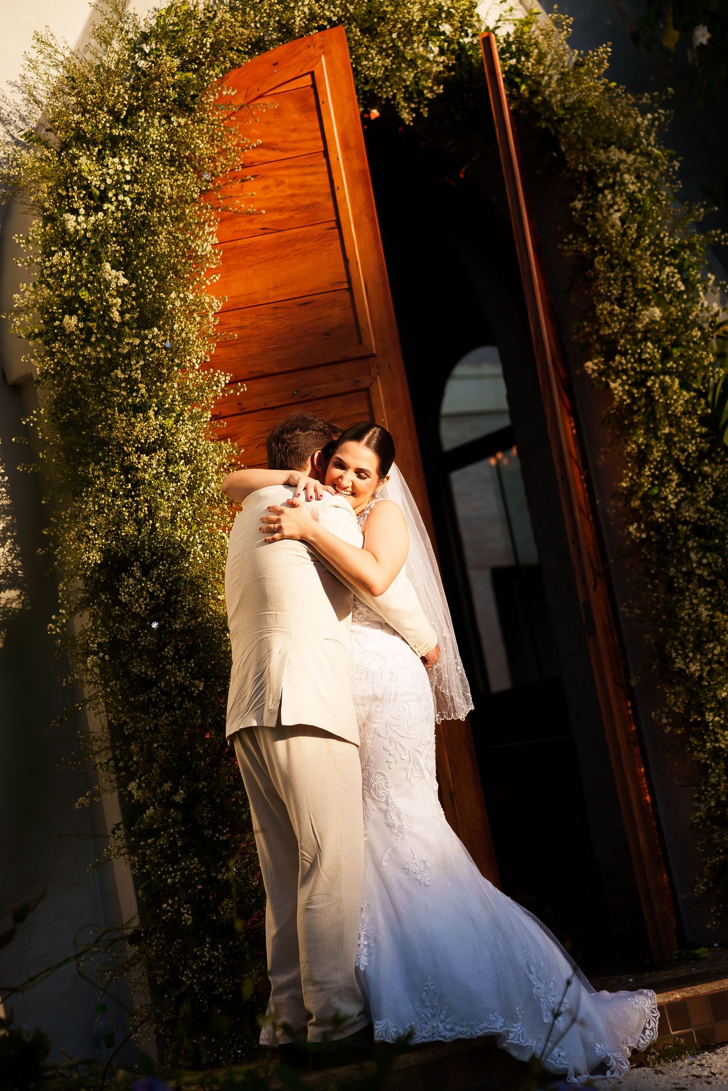 Bride and groom share a tender embrace outside wooden church doors adorned with lush greenery on their wedding day.