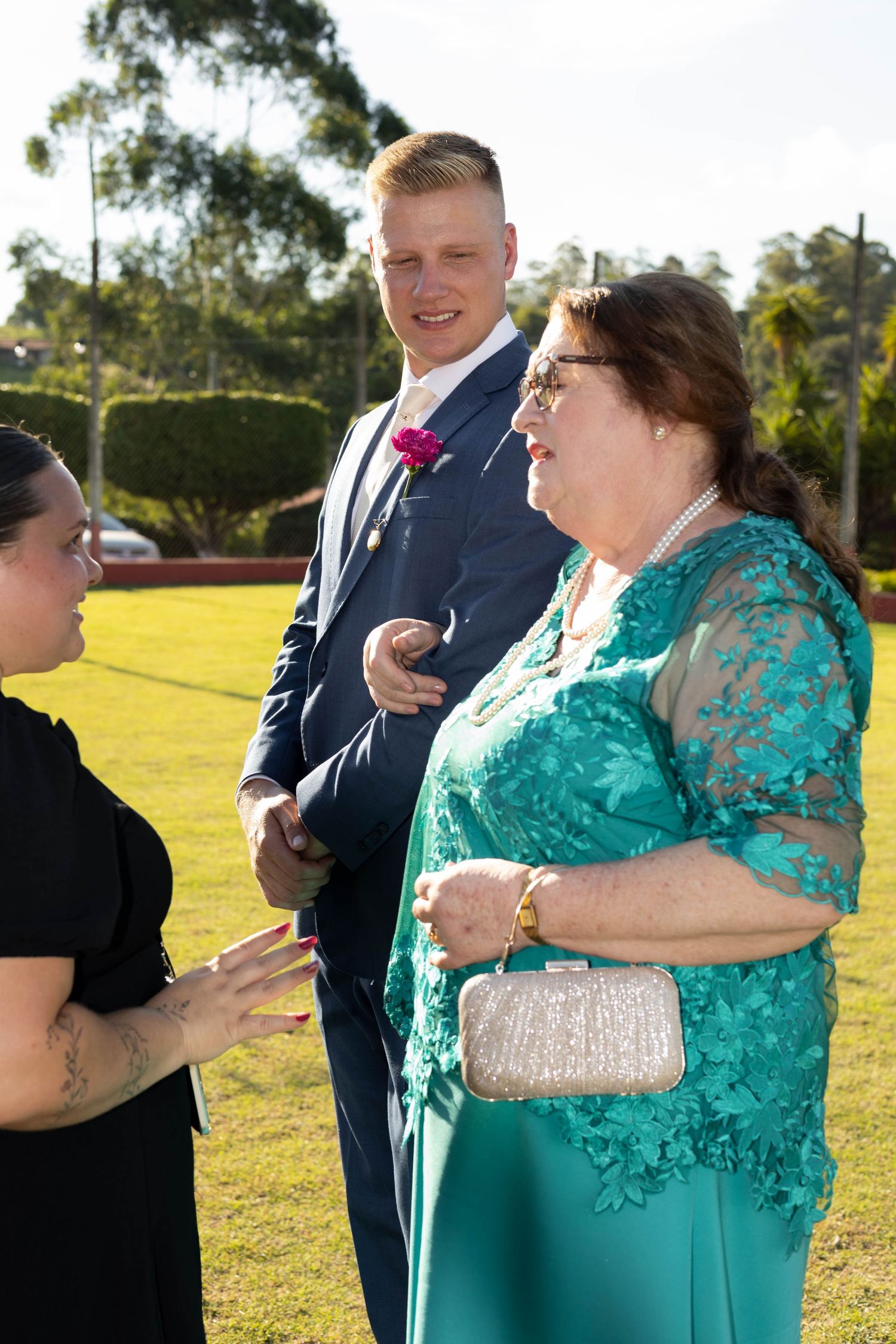 Man in navy suit with pink boutonniere and woman in teal lace dress conversing outdoors at a wedding reception.