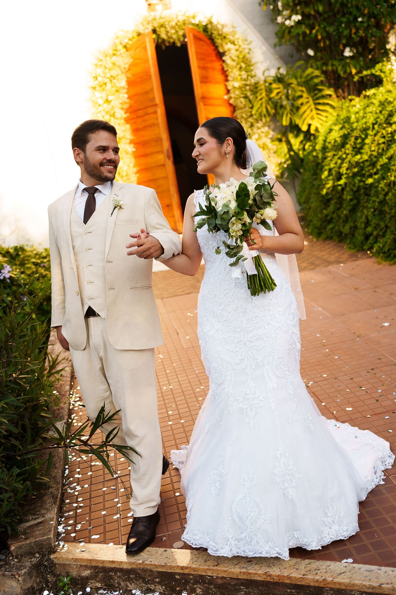 Happy newlyweds walking together, bride in white lace gown holding bouquet, groom in beige suit, outdoor garden venue.