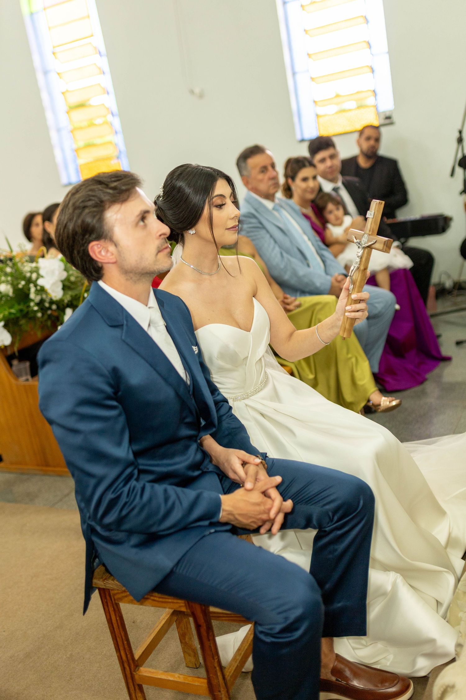 Bride holding crucifix and groom in blue suit seated at church wedding ceremony with guests in background.