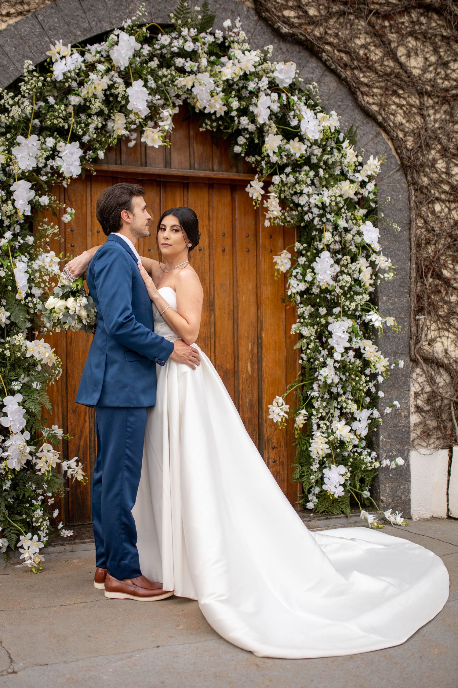 Bride and groom embrace before a floral arch with white flowers framing a wooden door on their wedding day.