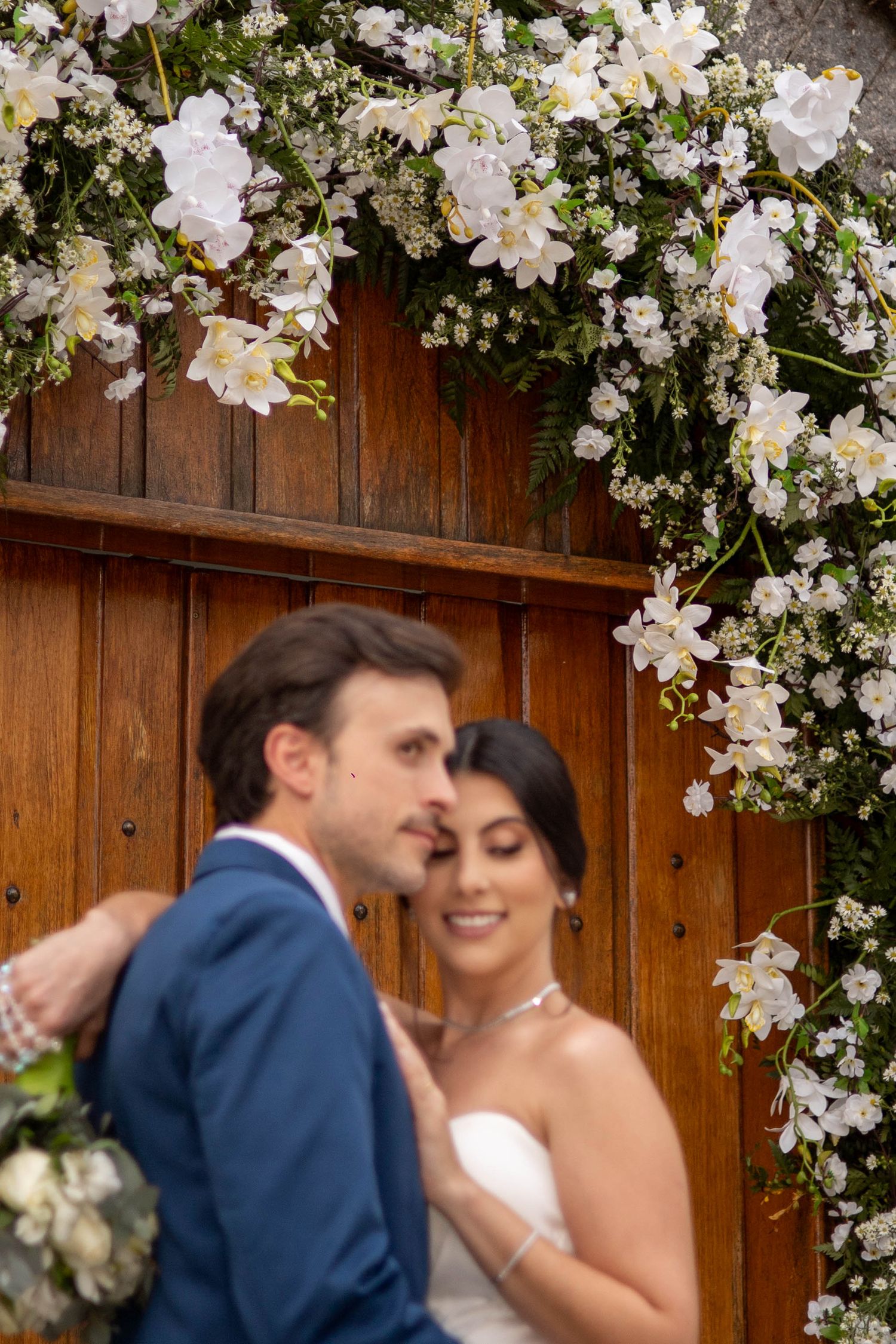 Bride and groom embrace under a floral arch of white blooms against a wooden door on their wedding day.