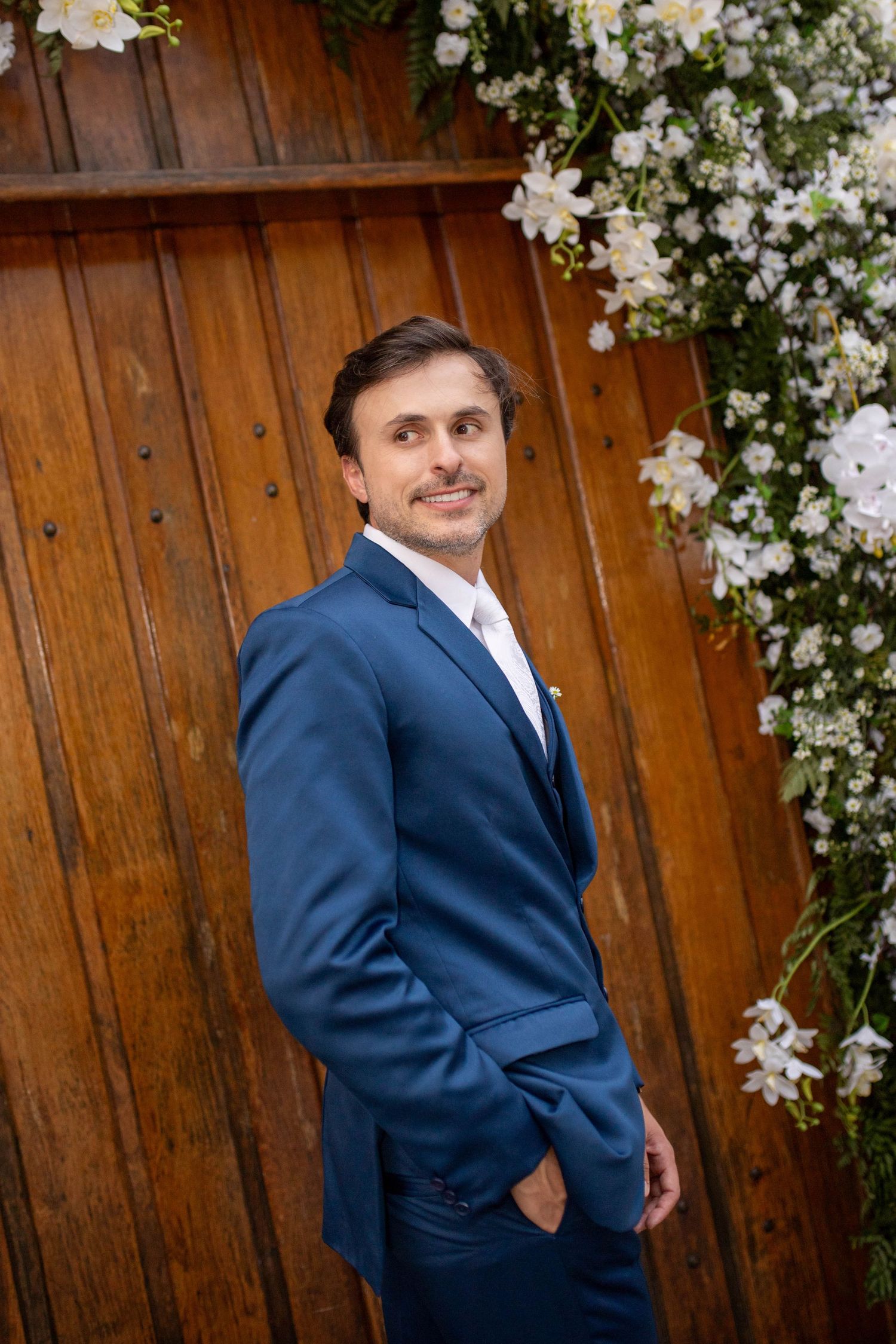 A groom in a blue suit smiles confidently beside a wooden backdrop adorned with white flowers.
