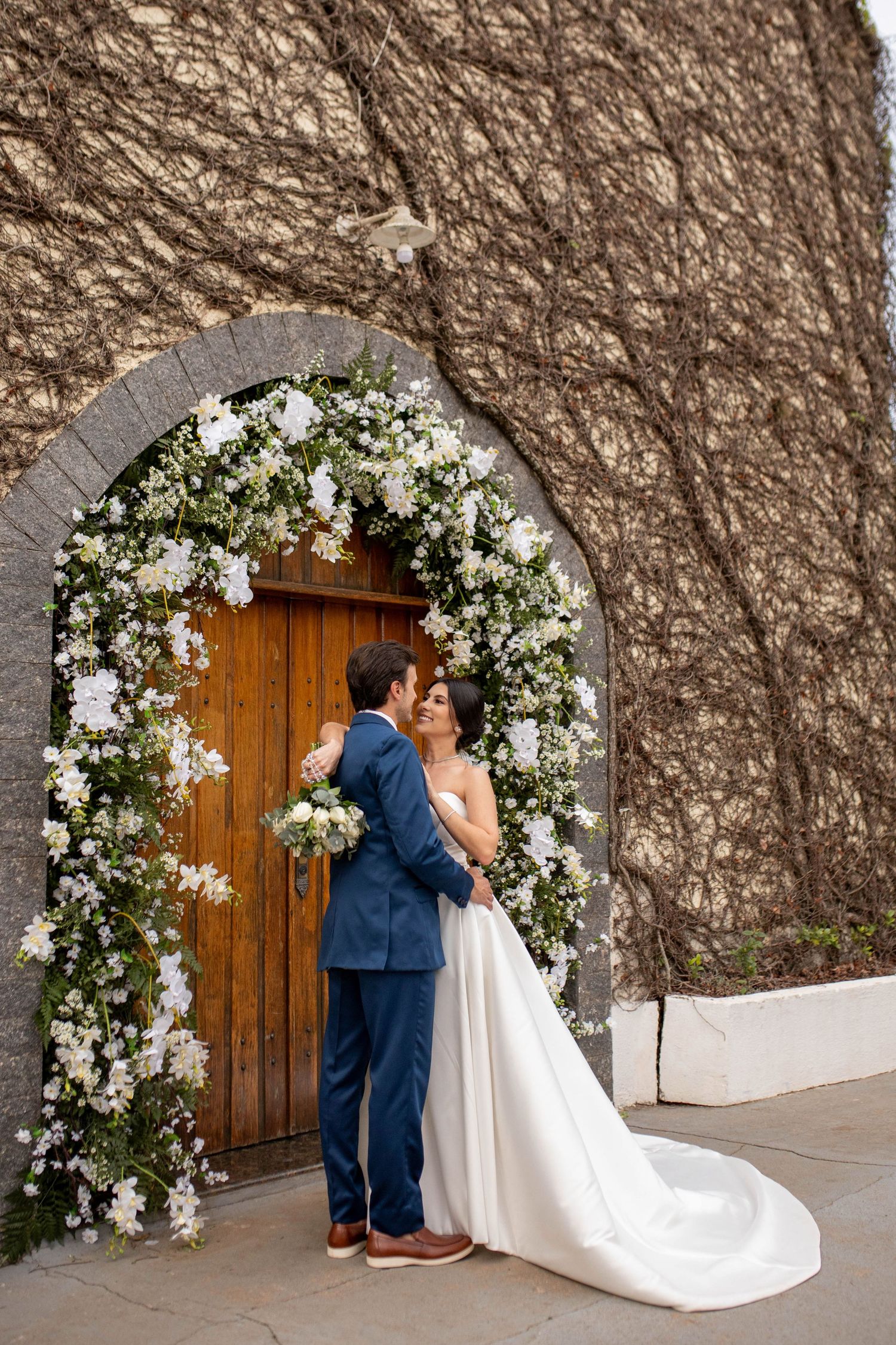 Bride and groom embrace under a white floral arch at a wooden door, surrounded by ivy-covered stone walls.