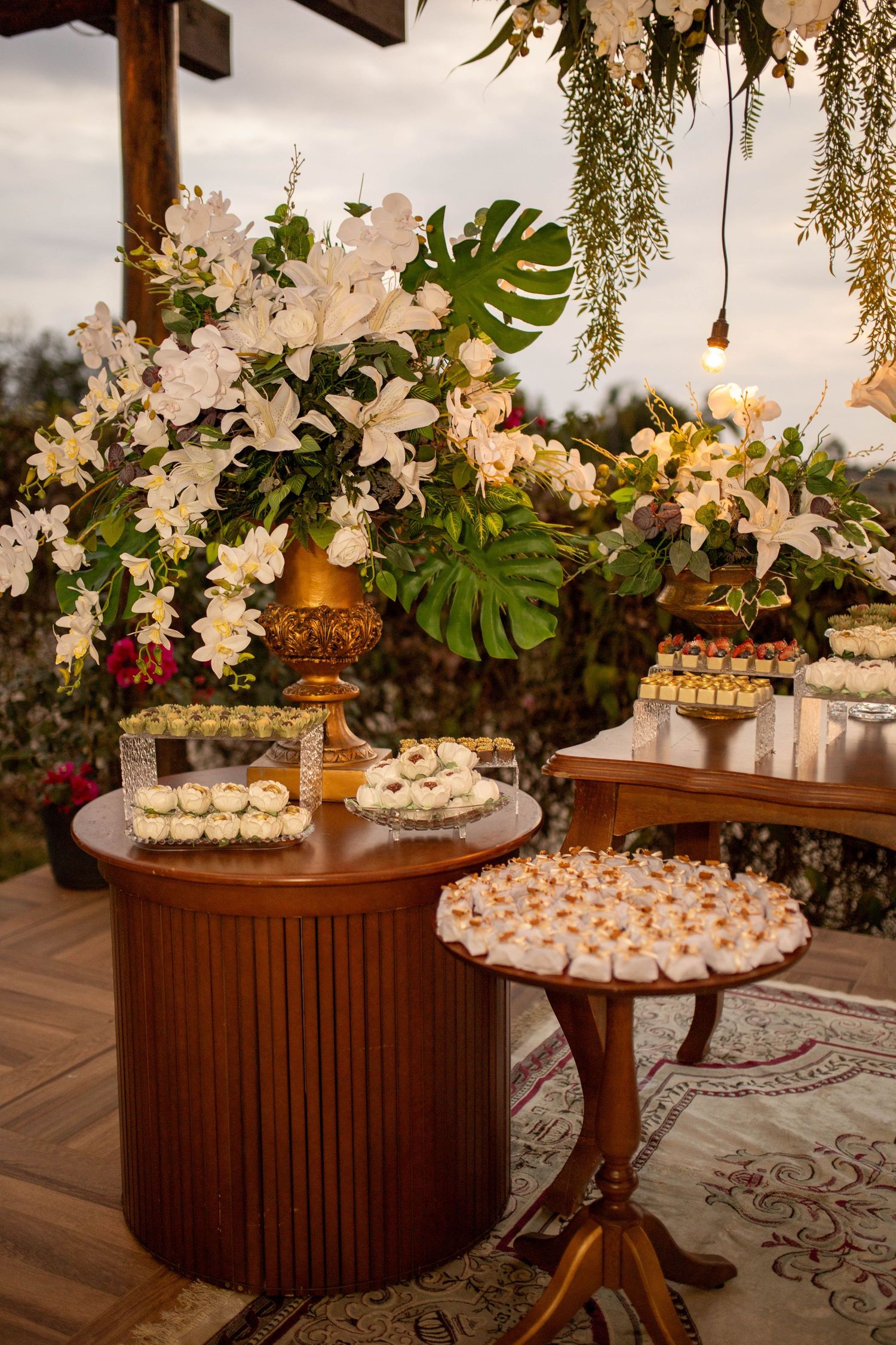 Elegant wedding dessert table with white floral arrangements, petit fours, and macarons on wooden tables under hanging greenery.