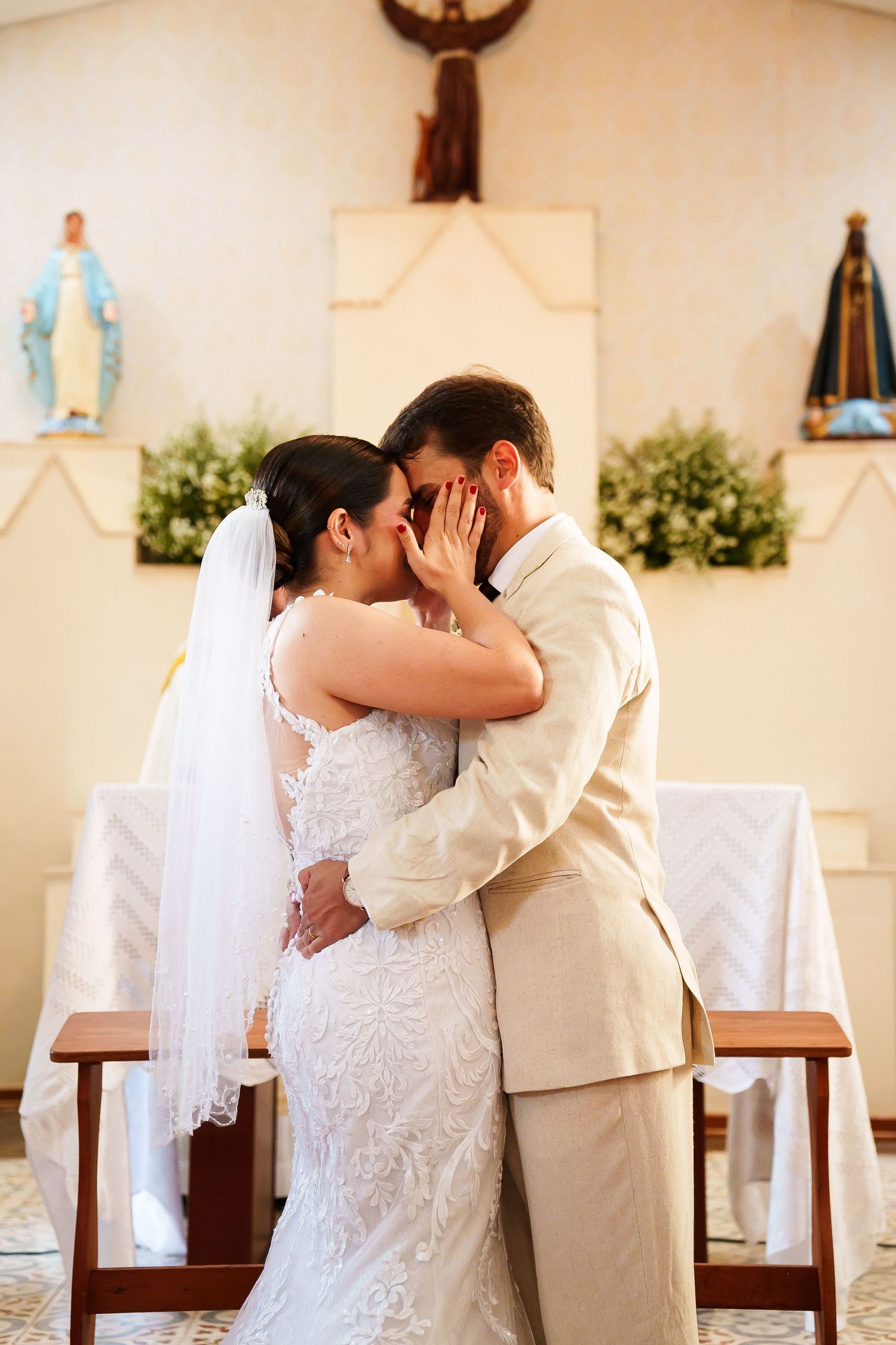 Bride and groom share first kiss at church altar, bride in lace gown with veil, groom in beige suit.