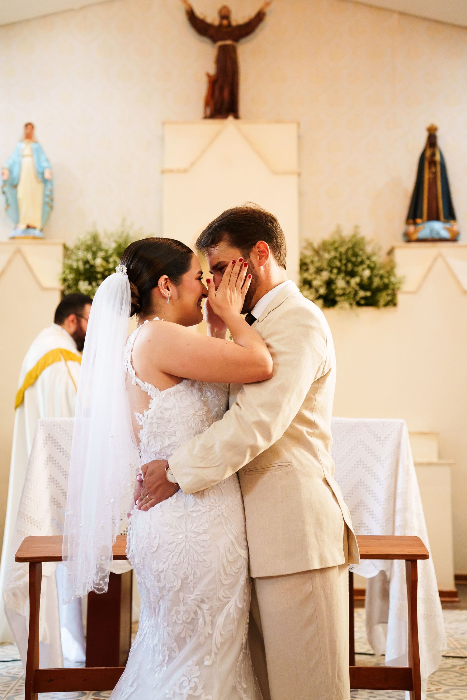 Bride and groom share first kiss at church altar during wedding ceremony, with priest and religious statues in background.