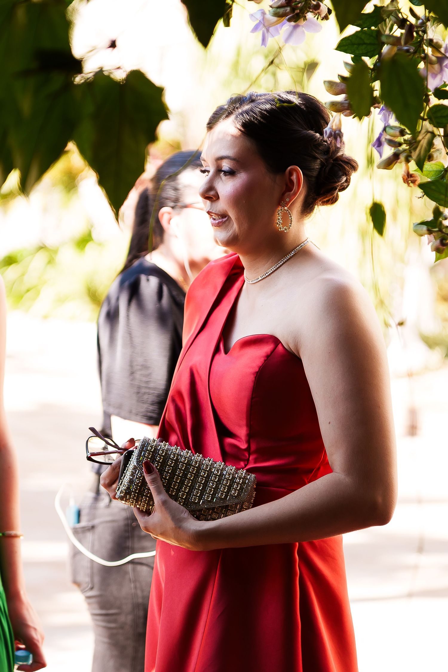 Woman in red strapless gown holding clutch purse at outdoor formal event surrounded by greenery.