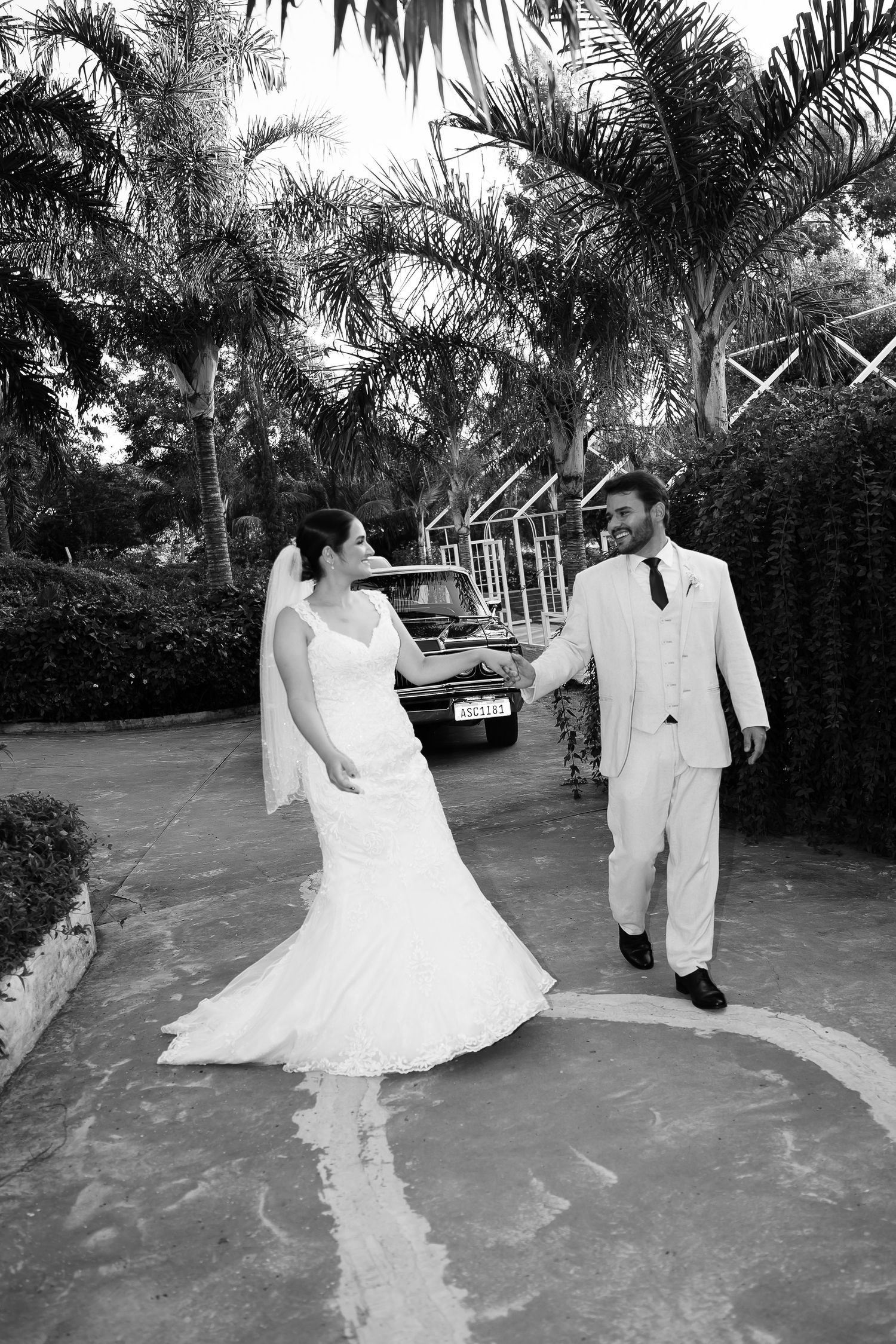 Black and white photo of a bride and groom walking hand in hand outdoors, with palm trees and a vintage car in the background.