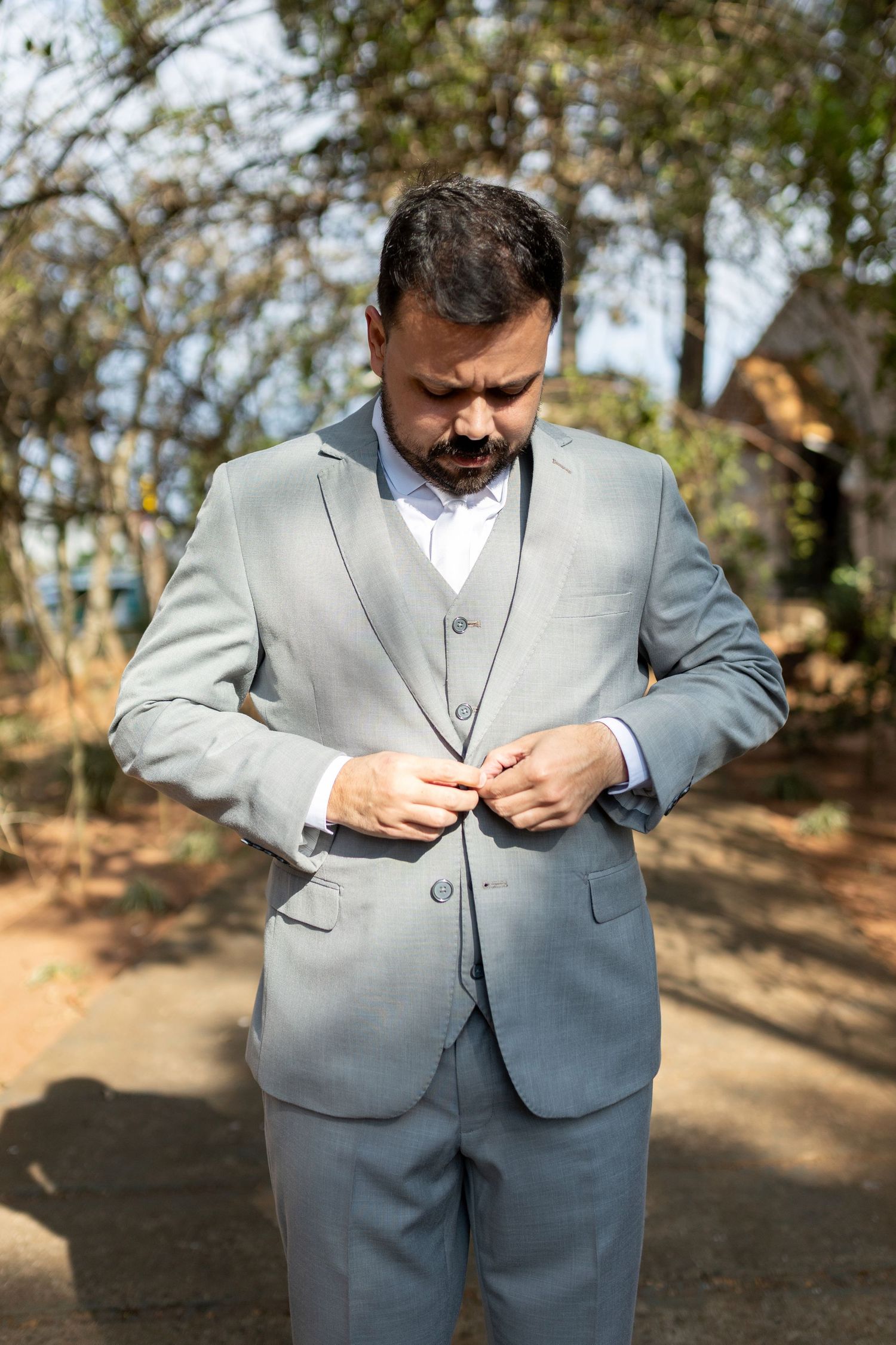 Man in gray three-piece suit buttoning jacket outdoors in a park setting.