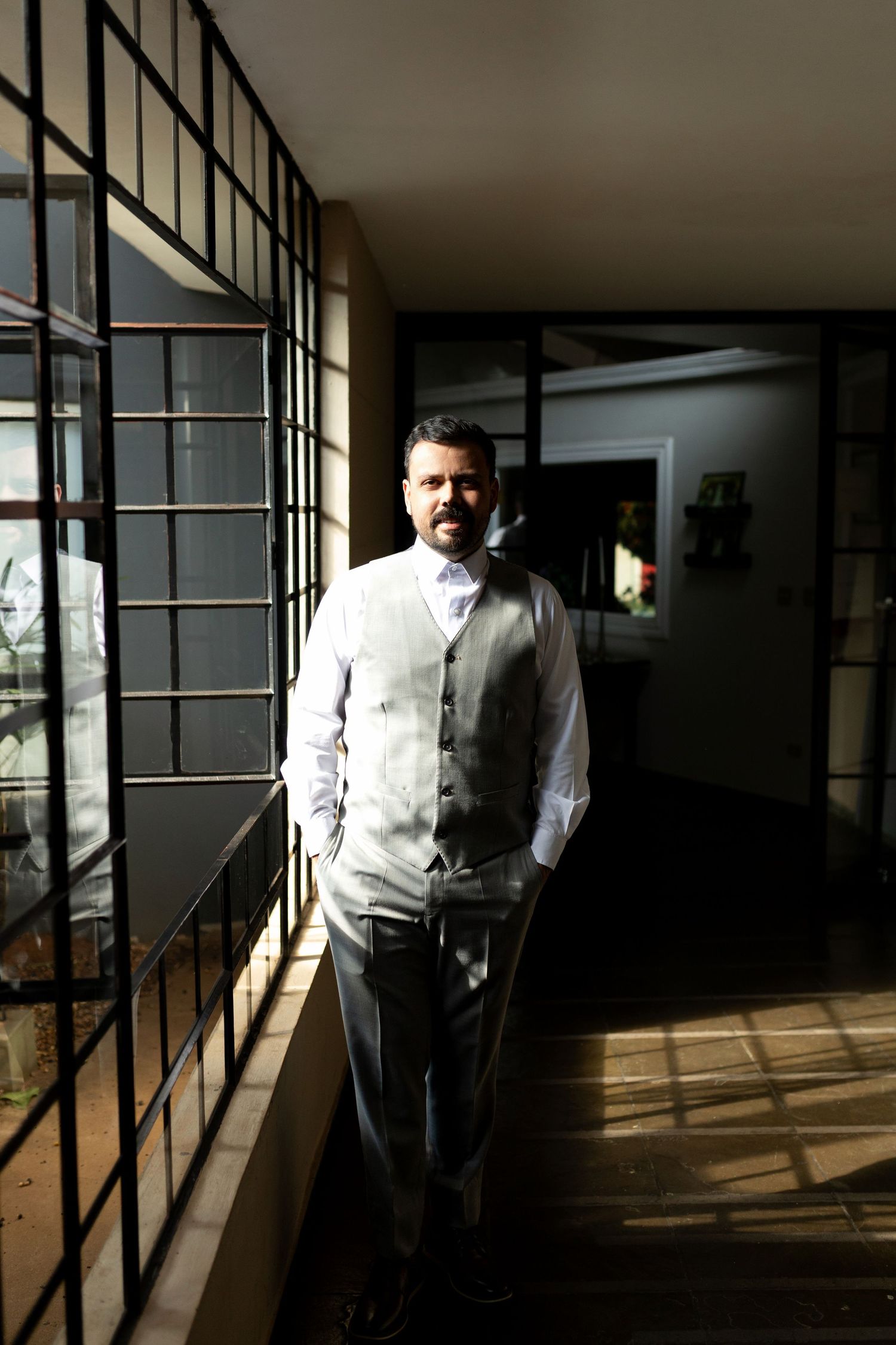 Man in gray vest and white shirt standing in sunlit hallway with metal-framed windows.
