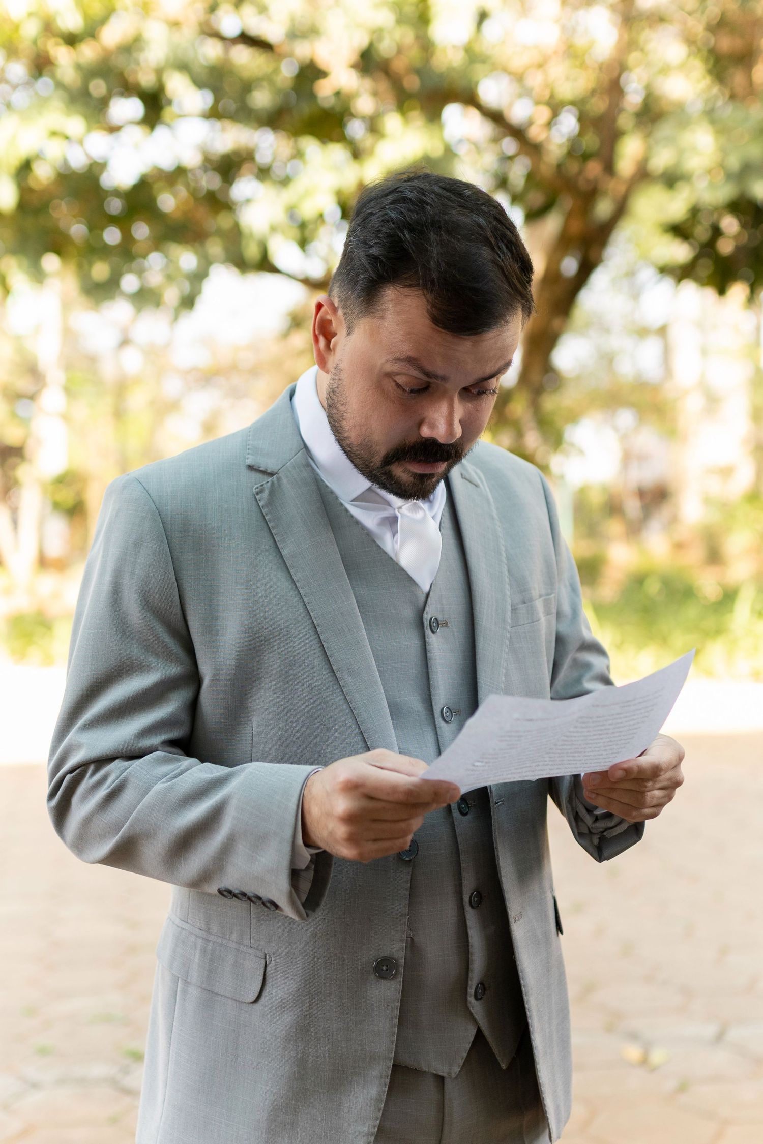 Elegant groom in a gray three-piece suit reading a paper document outdoors in a sunlit garden setting.