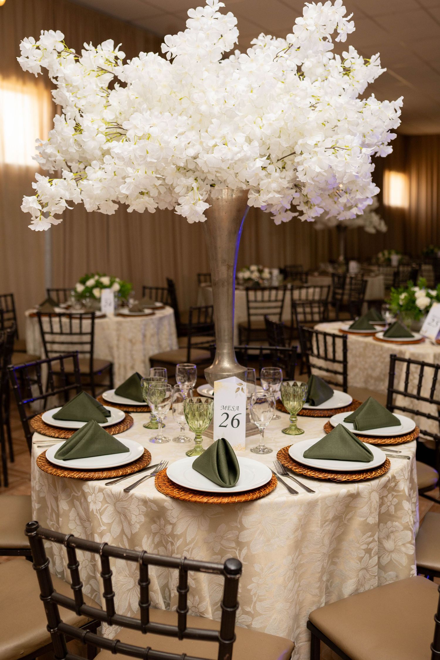 Elegant wedding reception table with tall white floral centerpiece, green napkins, gold chargers, and ivory linen in a banquet hall.