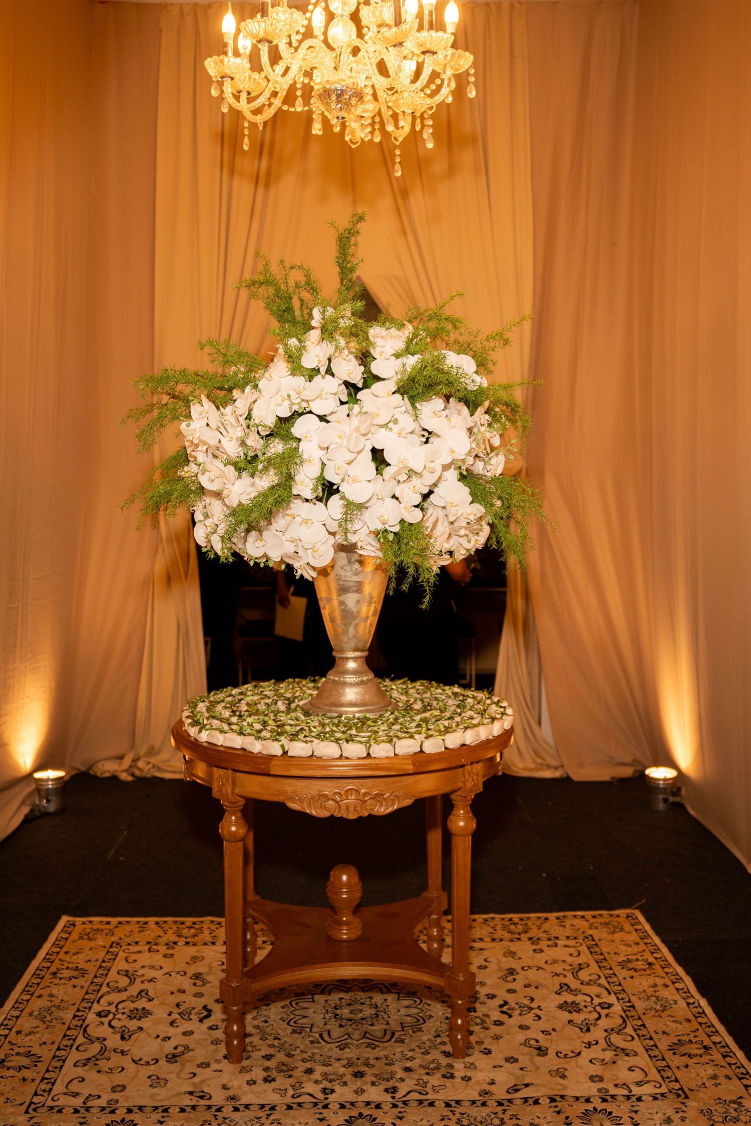 Elegant wooden table with white floral arrangement in silver vase, crystal chandelier, and draped fabric backdrop.