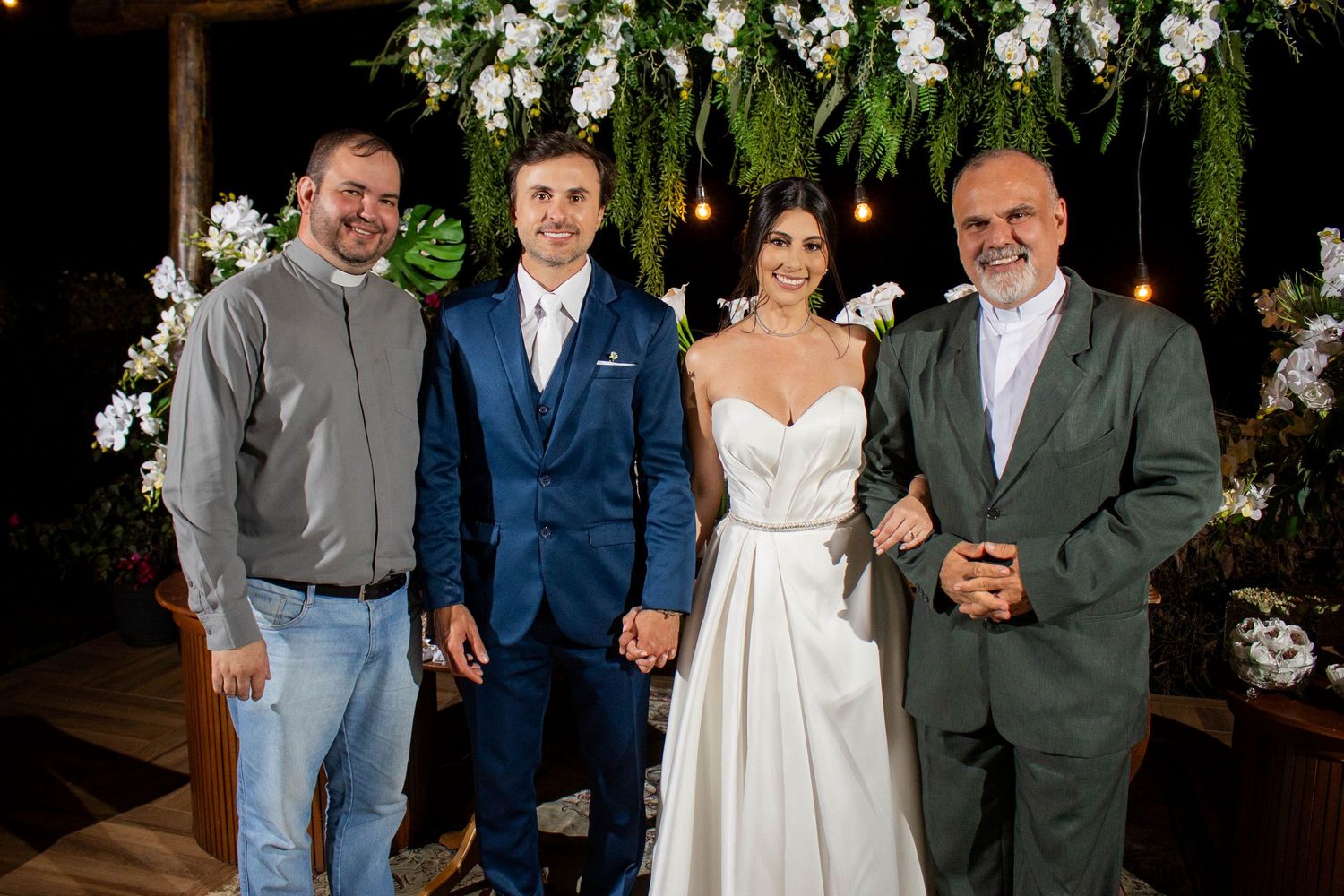 Bride and groom pose with two men at an elegant outdoor wedding ceremony decorated with white flowers and greenery.