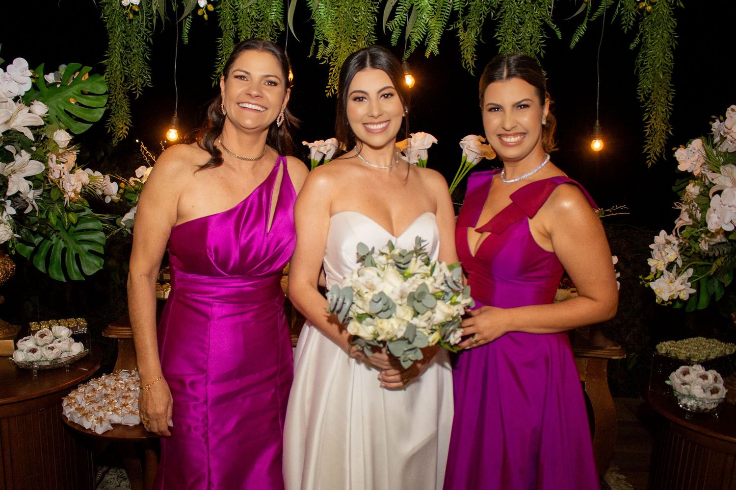 Bride in white gown holds bouquet flanked by two bridesmaids in fuchsia dresses at elegant floral wedding reception.