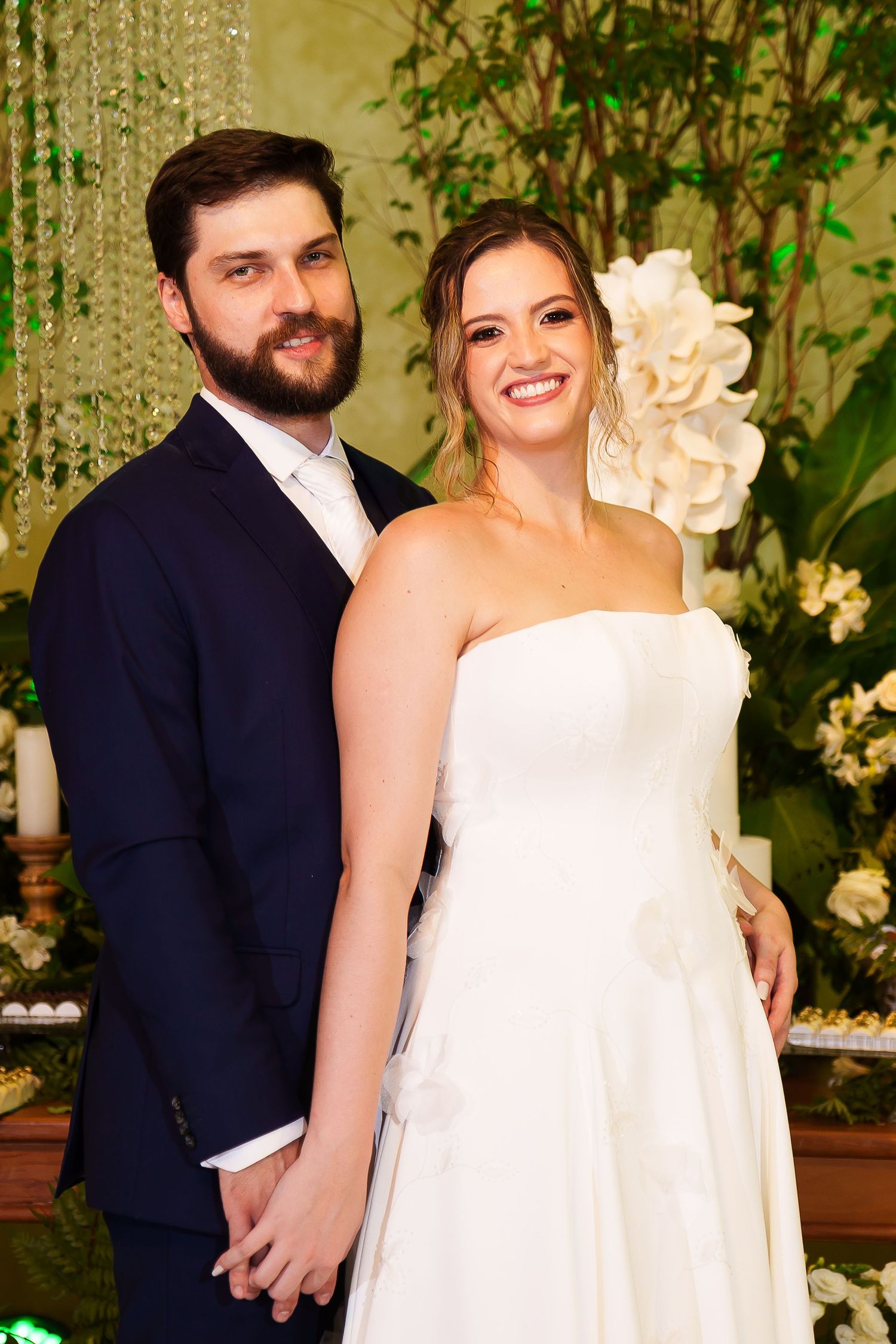 Smiling bride in white strapless gown and groom in navy suit hold hands at elegant floral wedding reception.