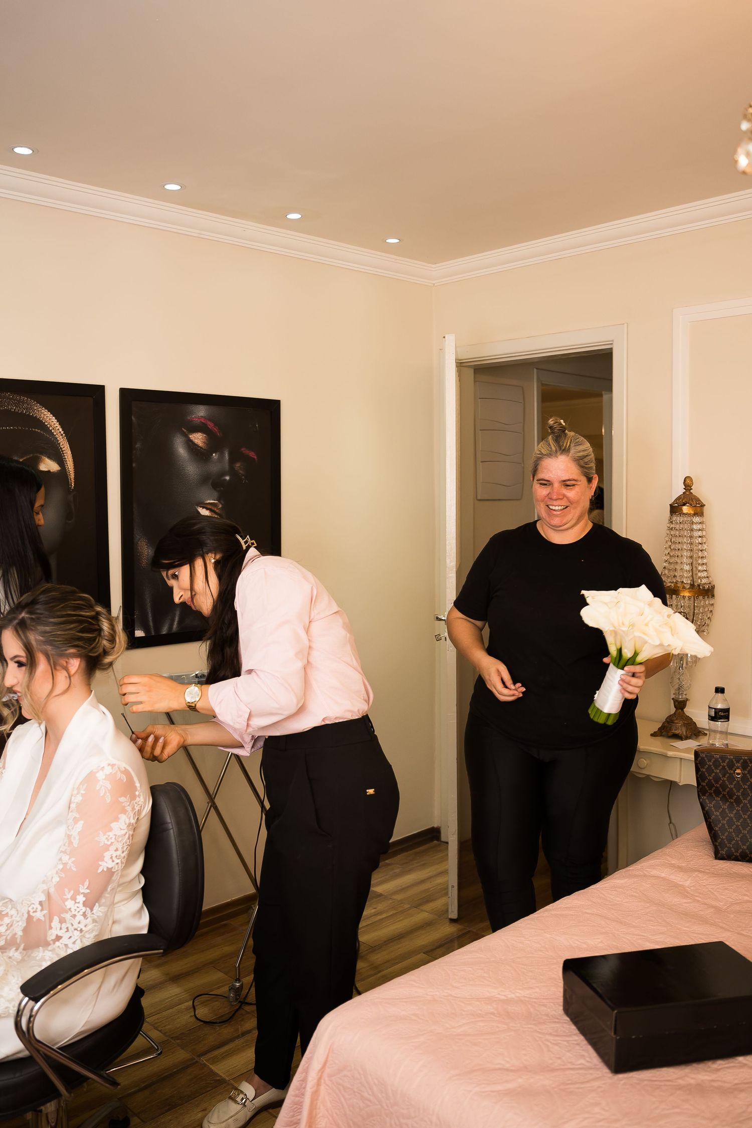 Bride getting hair styled by beautician while woman in black holds white calla lily bouquet in bridal suite.