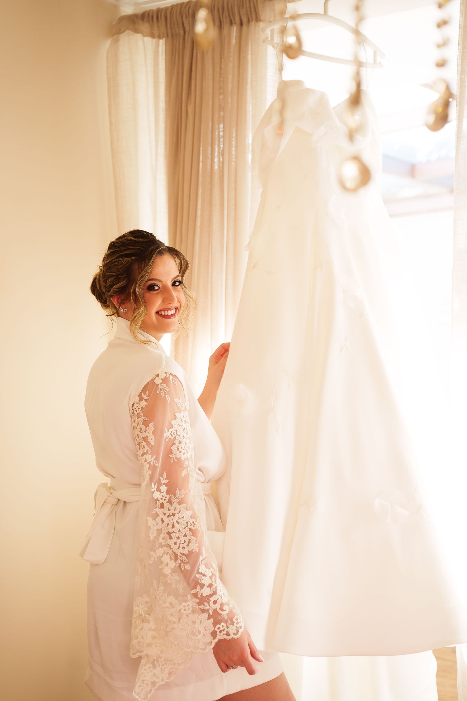 Smiling bride in lace robe holding her white wedding dress on a hanger in a softly lit bridal suite.