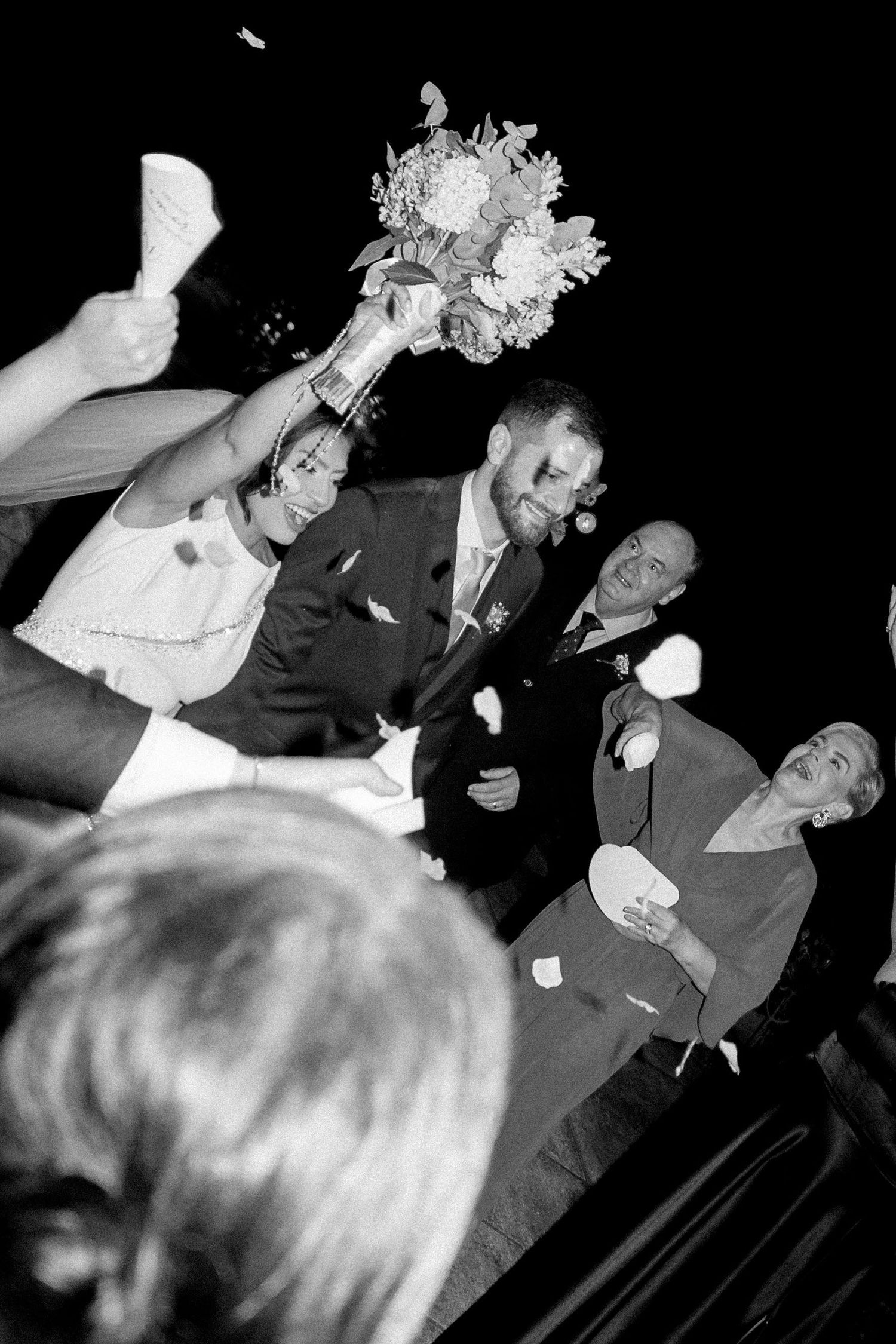 Bride raises bouquet joyfully as groom and guests celebrate at a wedding reception in black and white.