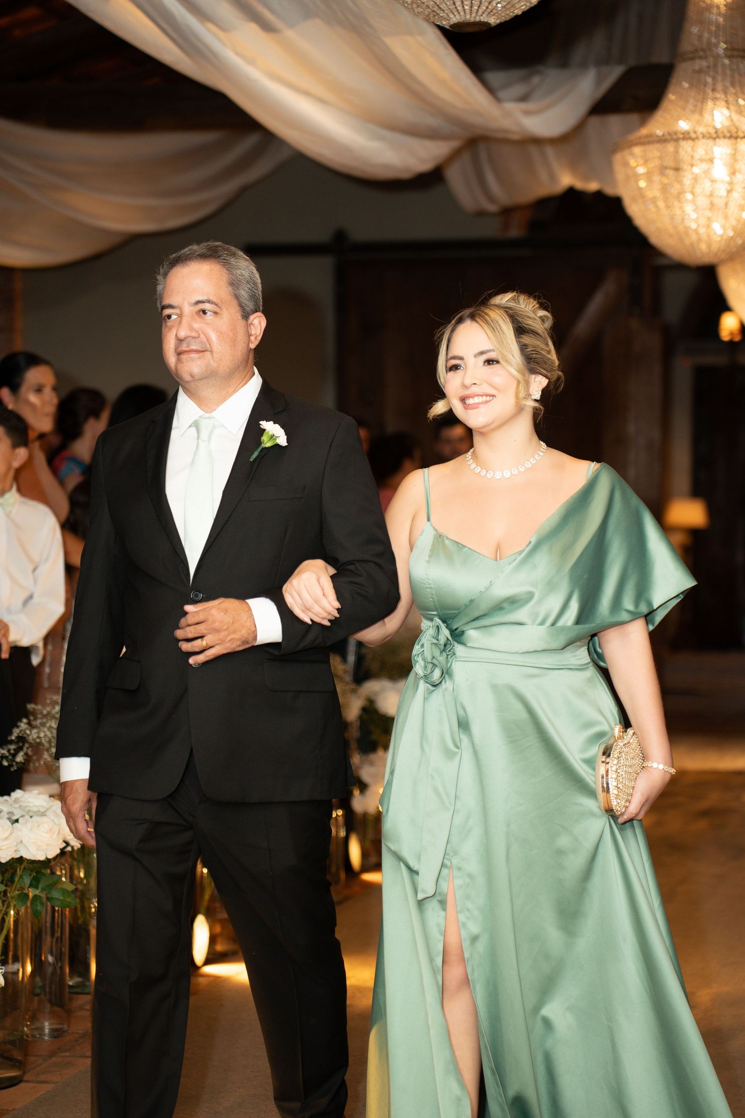 Elegant couple walking at wedding reception, man in tuxedo and woman in sage green satin gown with floral accessories.
