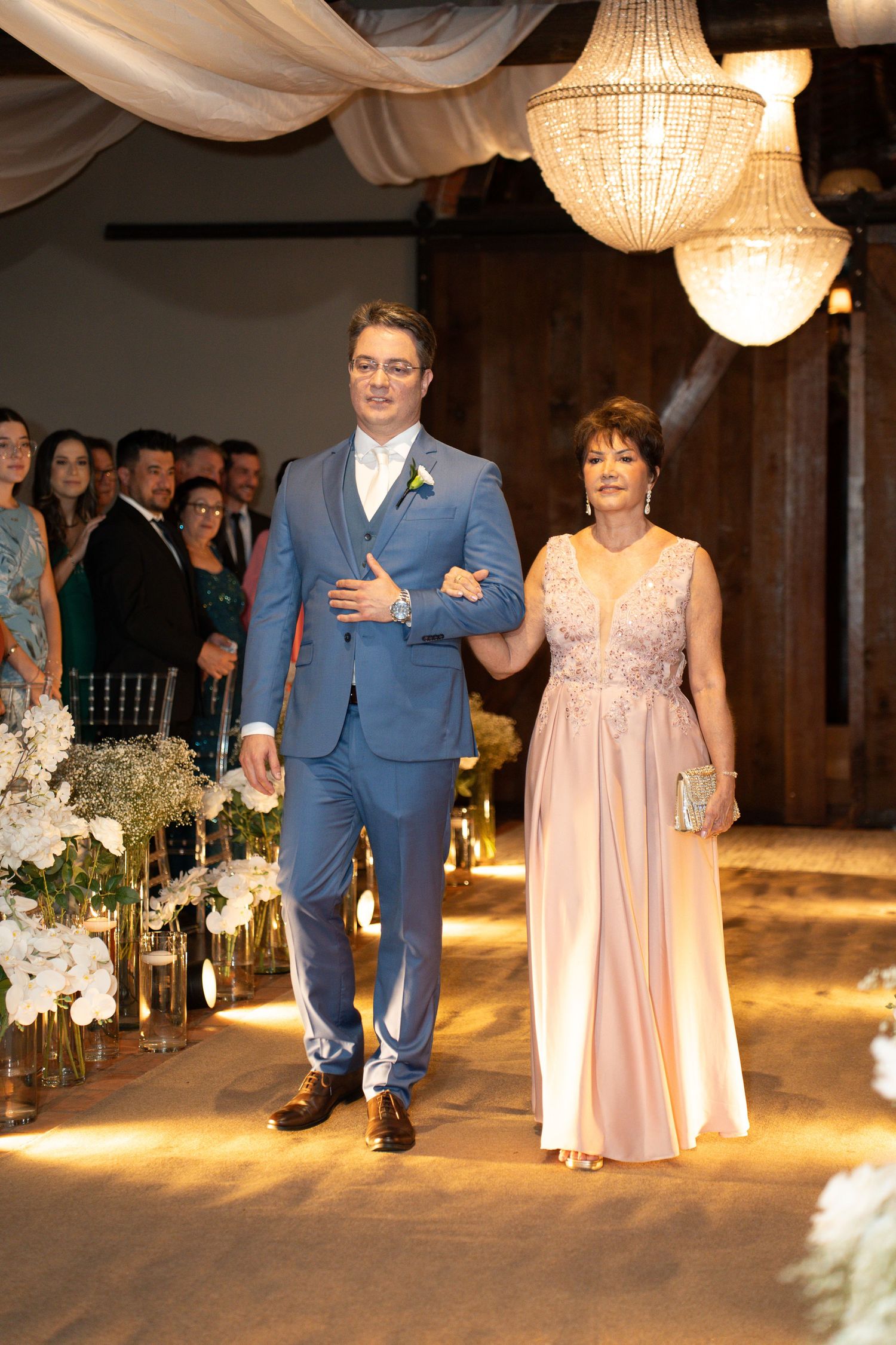 Groom in blue suit walks down wedding aisle with elegantly dressed woman in pink gown at indoor ceremony.