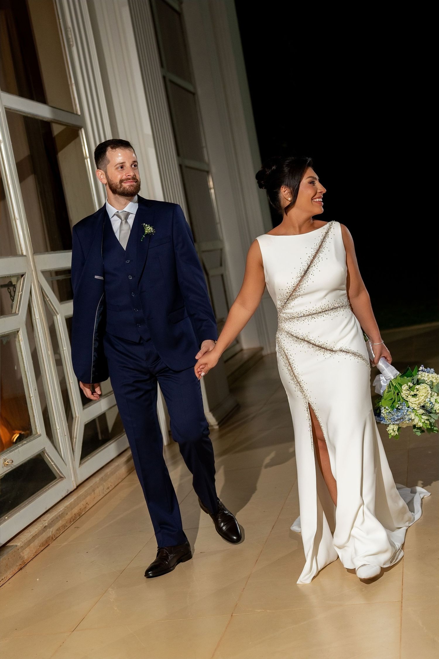 Bride in white gown and groom in navy suit walk hand in hand at night during their wedding reception.
